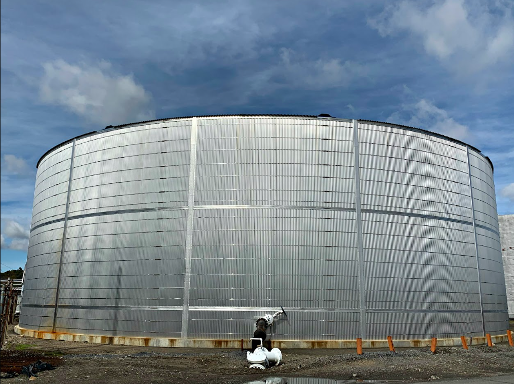 Large, cylindrical metal tank or silo with a curved roof, situated outdoors under a partly cloudy sky.