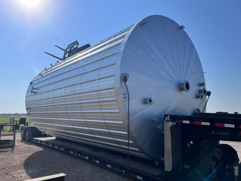 Large cylindrical tank on a flatbed trailer in an open field under a clear blue sky.