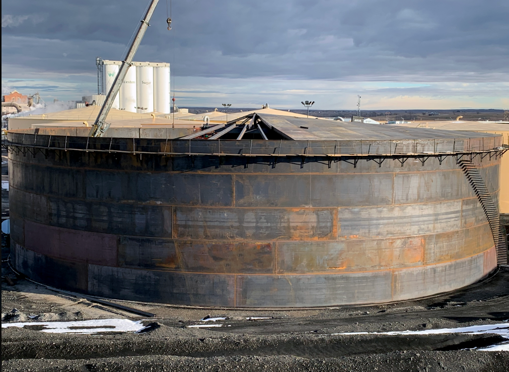 Large industrial storage tank with a staircase on the side, under a cloudy sky, with construction or maintenance equipment nearby.