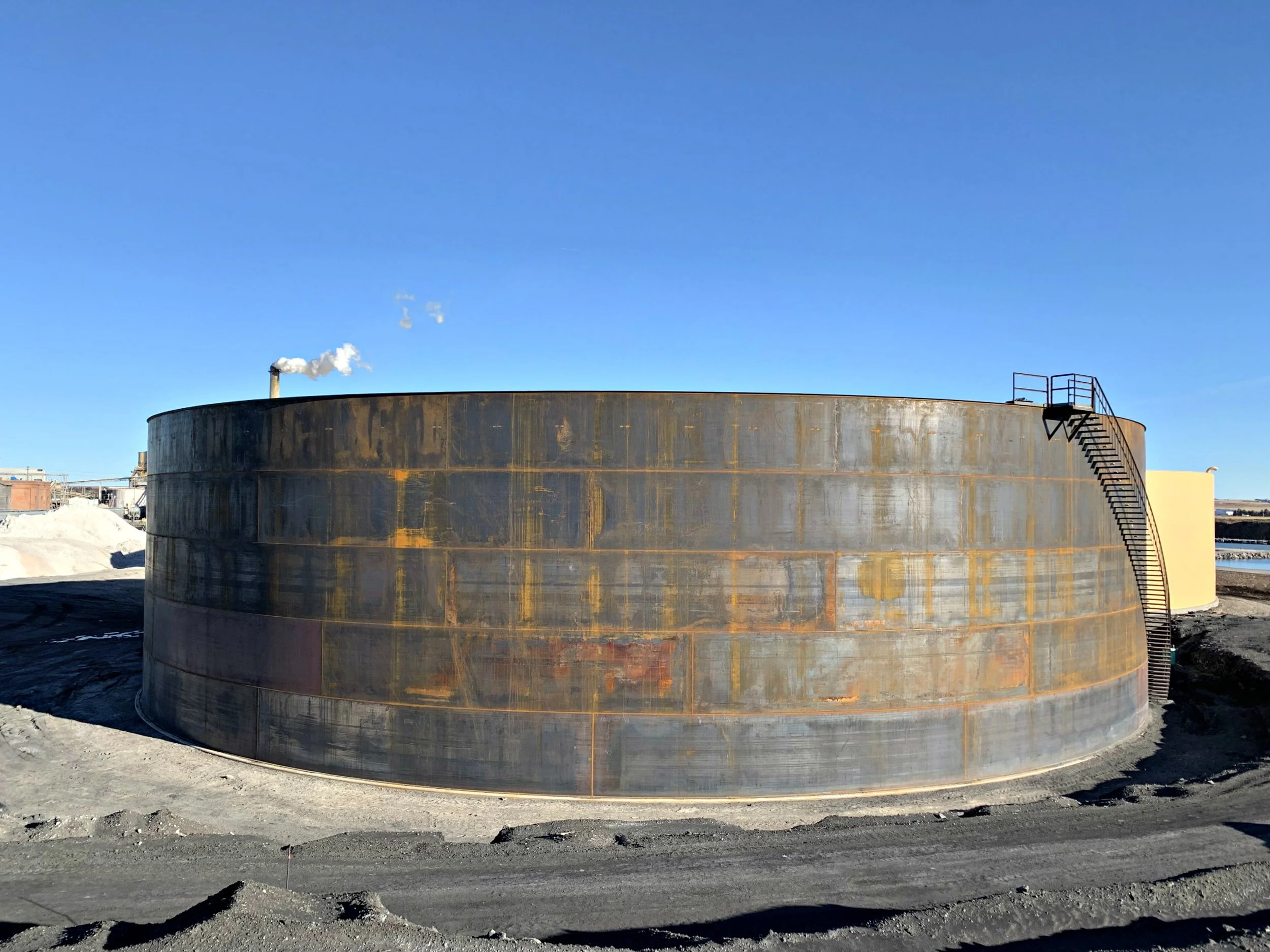 Large cylindrical water storage tank made of metal with visible rust and weathering, situated outdoors under a clear blue sky. There is a staircase on the right side providing access to the top of the tank.