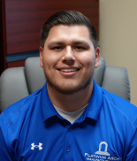 A young man with short dark hair, smiling, wearing a blue collared shirt, sitting in a gray office chair.