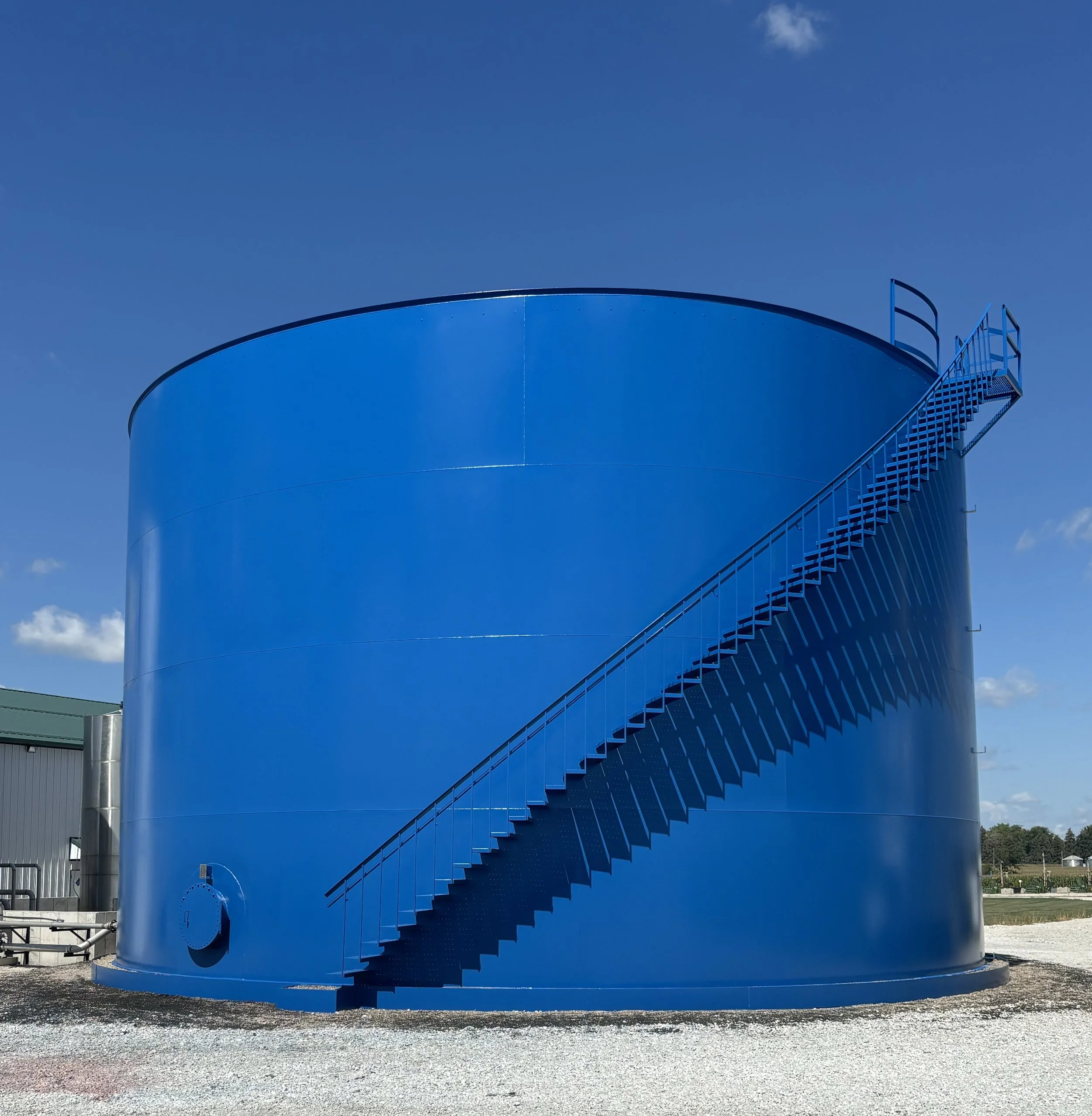 A large, shiny blue industrial storage tank with a spiral staircase on the side, set outdoors under a blue sky with some clouds.