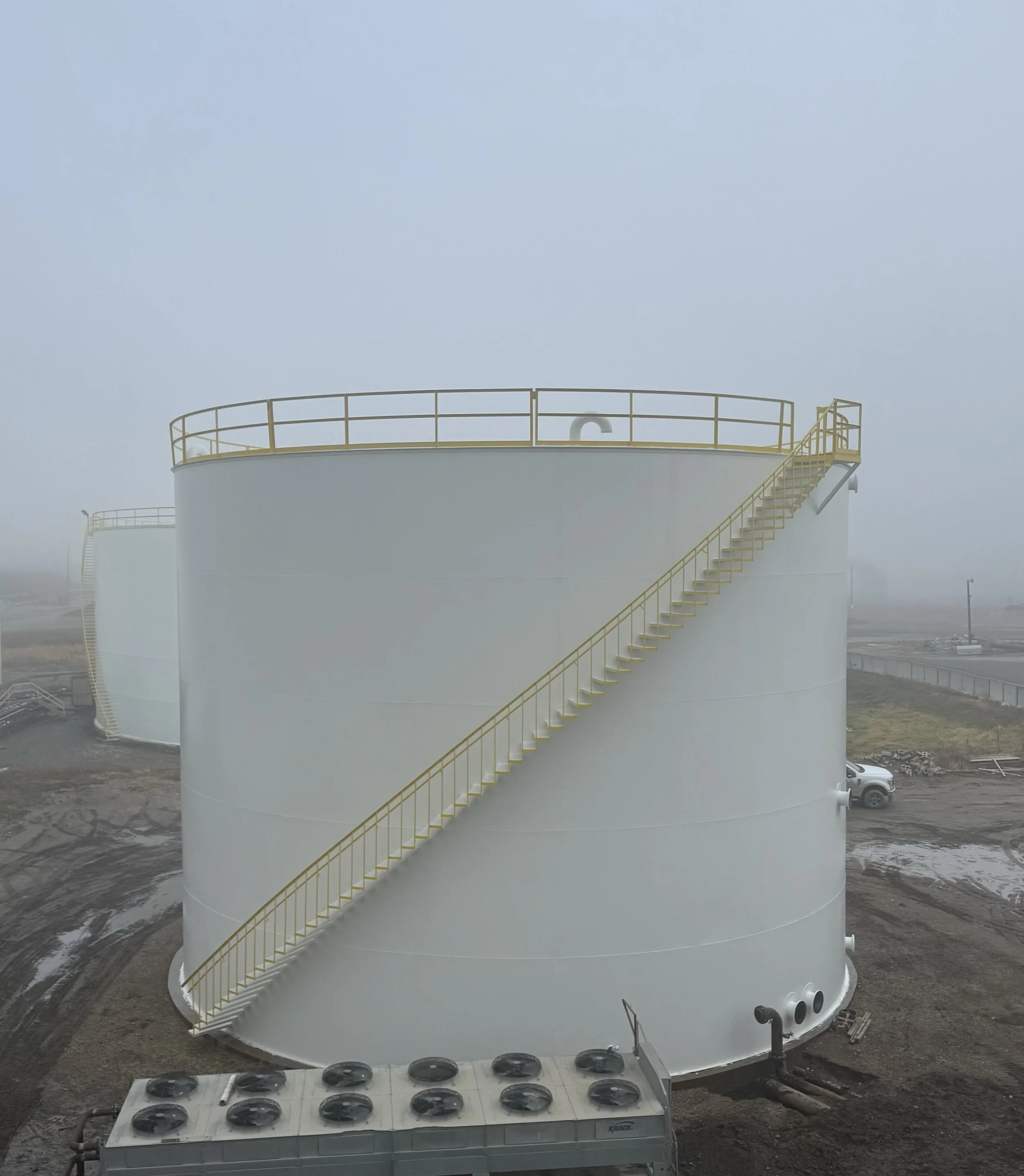Large white industrial tank with a yellow staircase and railing on its side, set outdoors in foggy weather, with industrial equipment and a truck in the background.