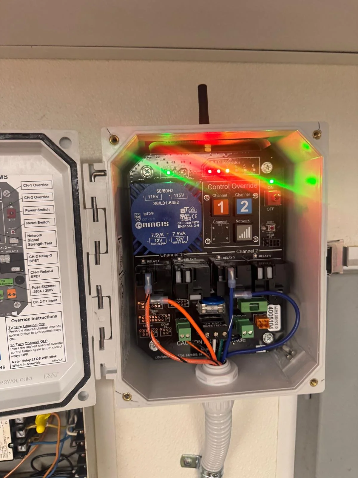 An electrical control panel containing a blue AMGIS relay, LED indicators, wiring, and control switches.