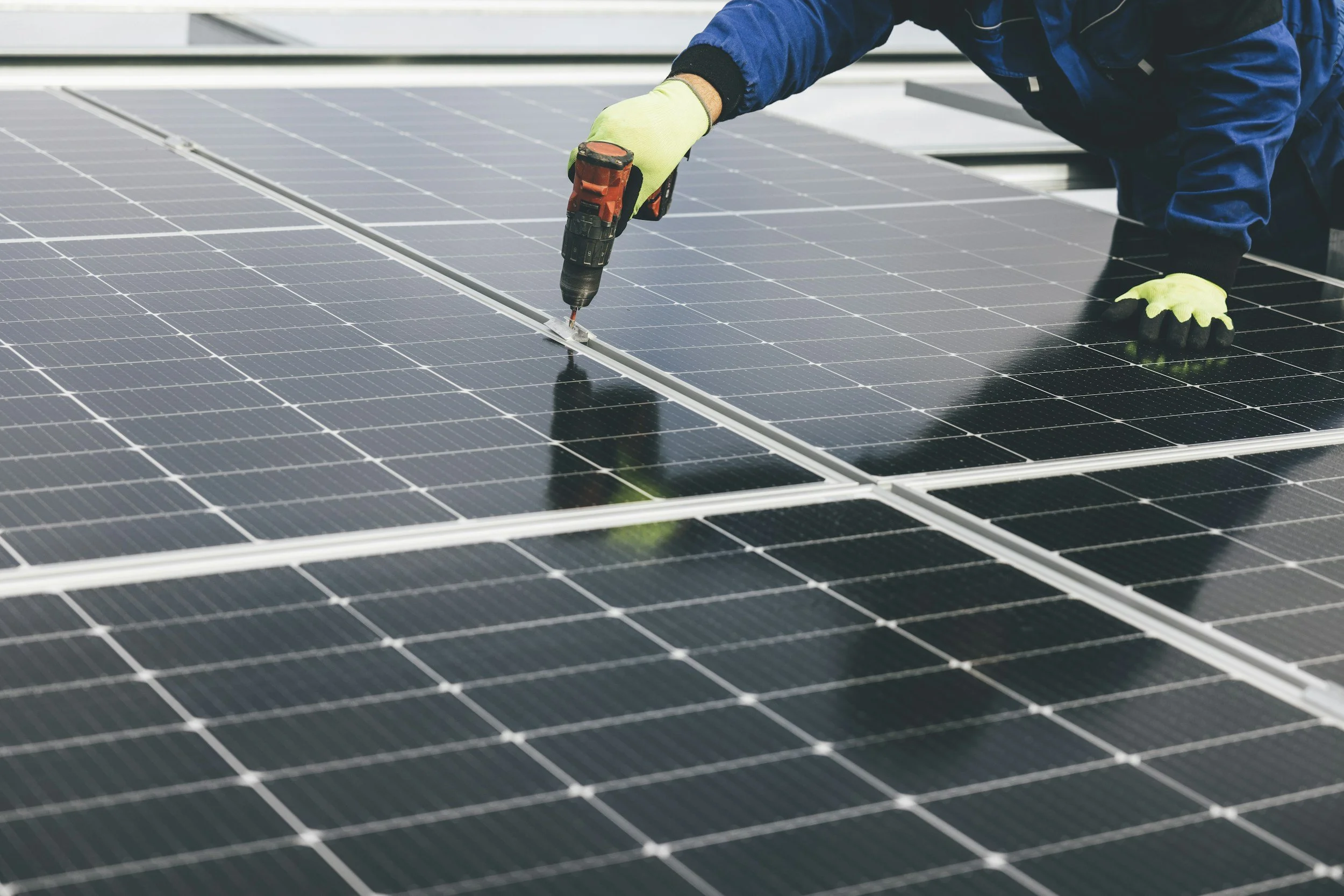 Worker installing or maintaining solar panels, wearing gloves and using a power drill.