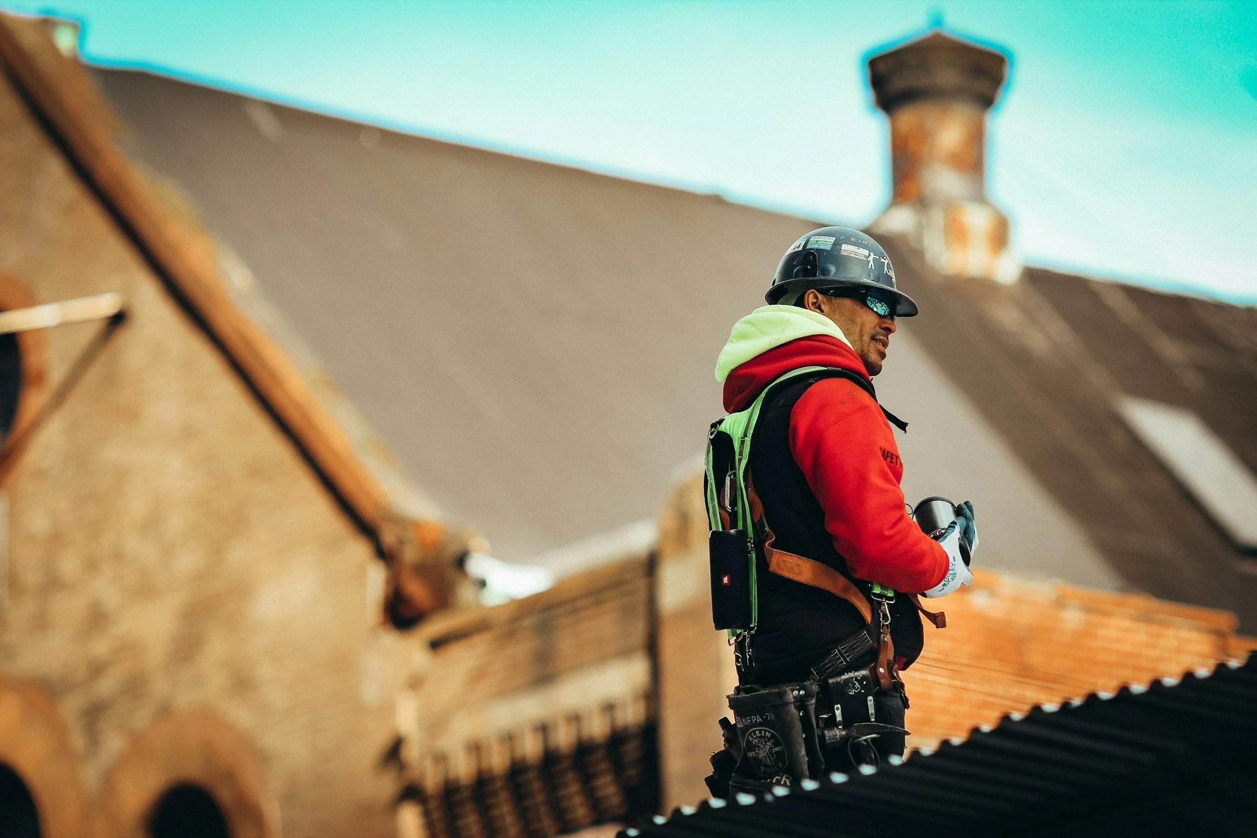 A construction worker in a red and green safety jacket, black safety helmet, and sunglasses standing among wooden structures during construction.
