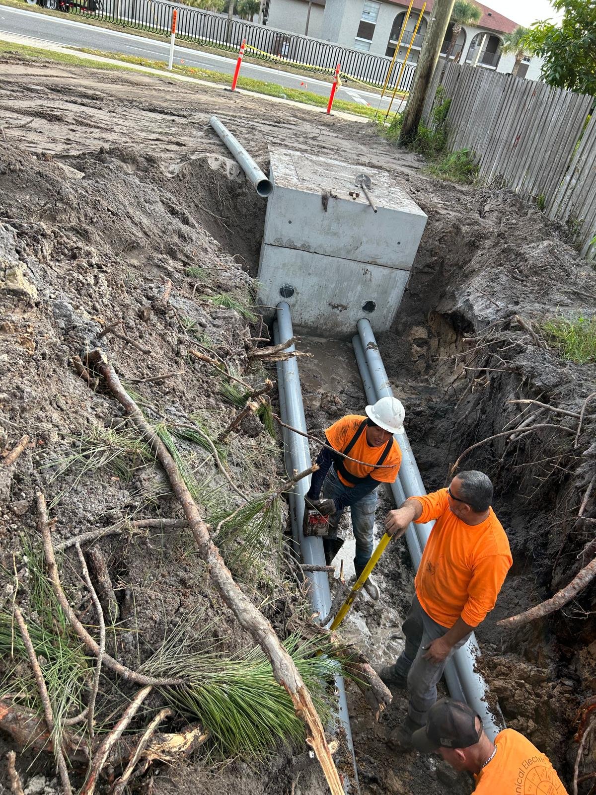 Workers installing underground utility pipes in a trench next to a street with houses and trees in the background.