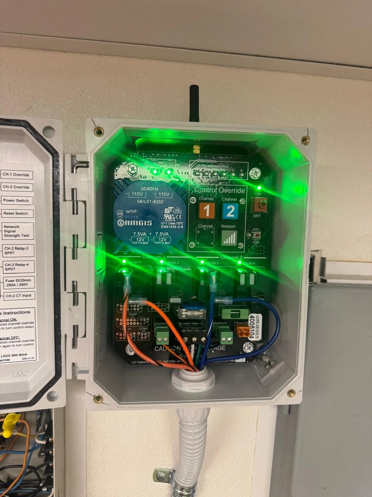 An electrical control panel with green LED lights and various wires and electronic components inside a metal enclosure. A label on the left lists switches and test functions.