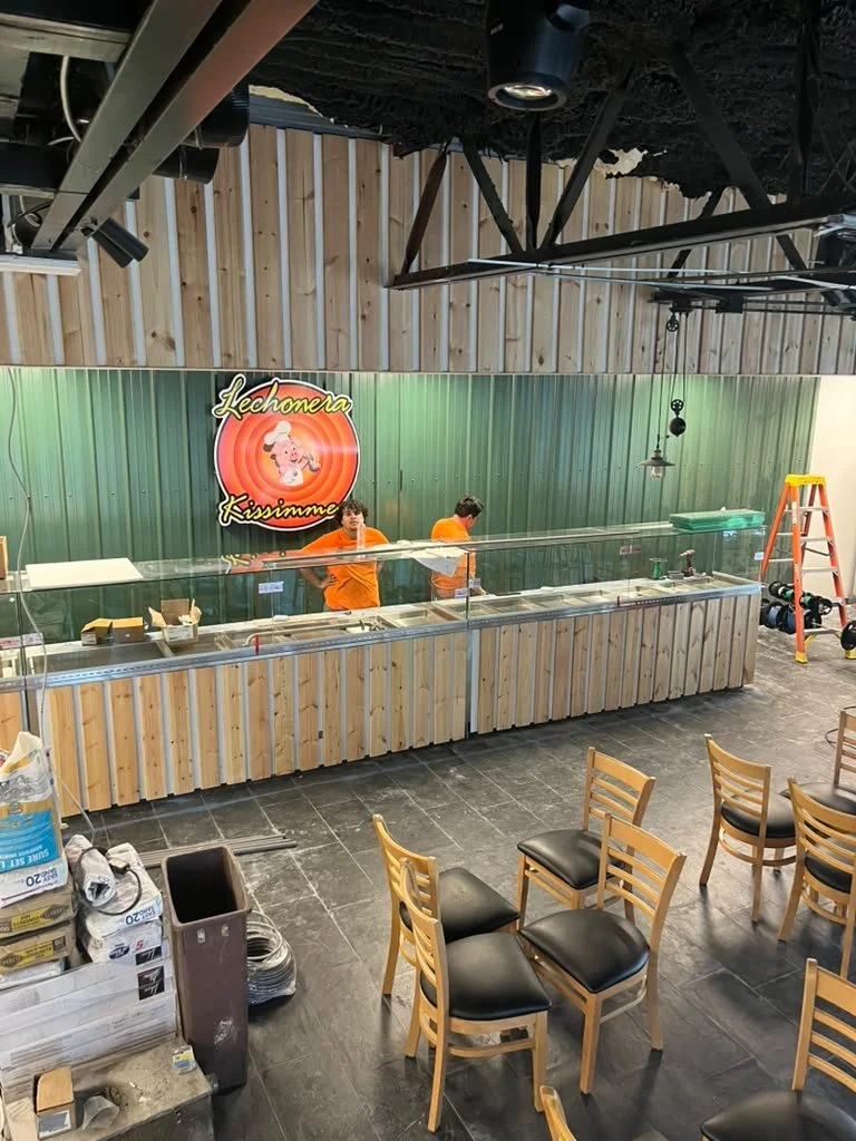 Inside a restaurant or food service area with two workers behind a glass counter, a green wall with a colorful sign reading 'Lechonera Kissimme,' and wooden chairs arranged in front. There is construction or setup equipment like a ladder and tools nearby.