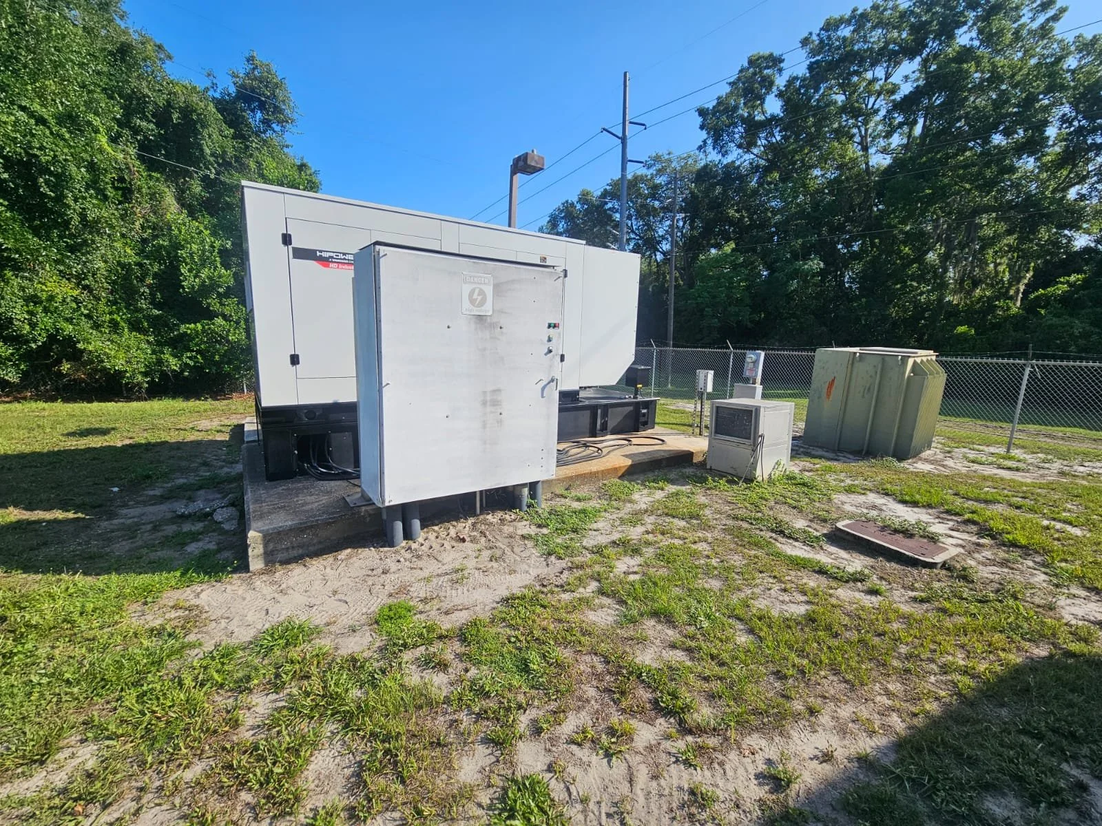 An outdoor electrical substation with transformers and electrical equipment, surrounded by grass, trees, and a chain-link fence.