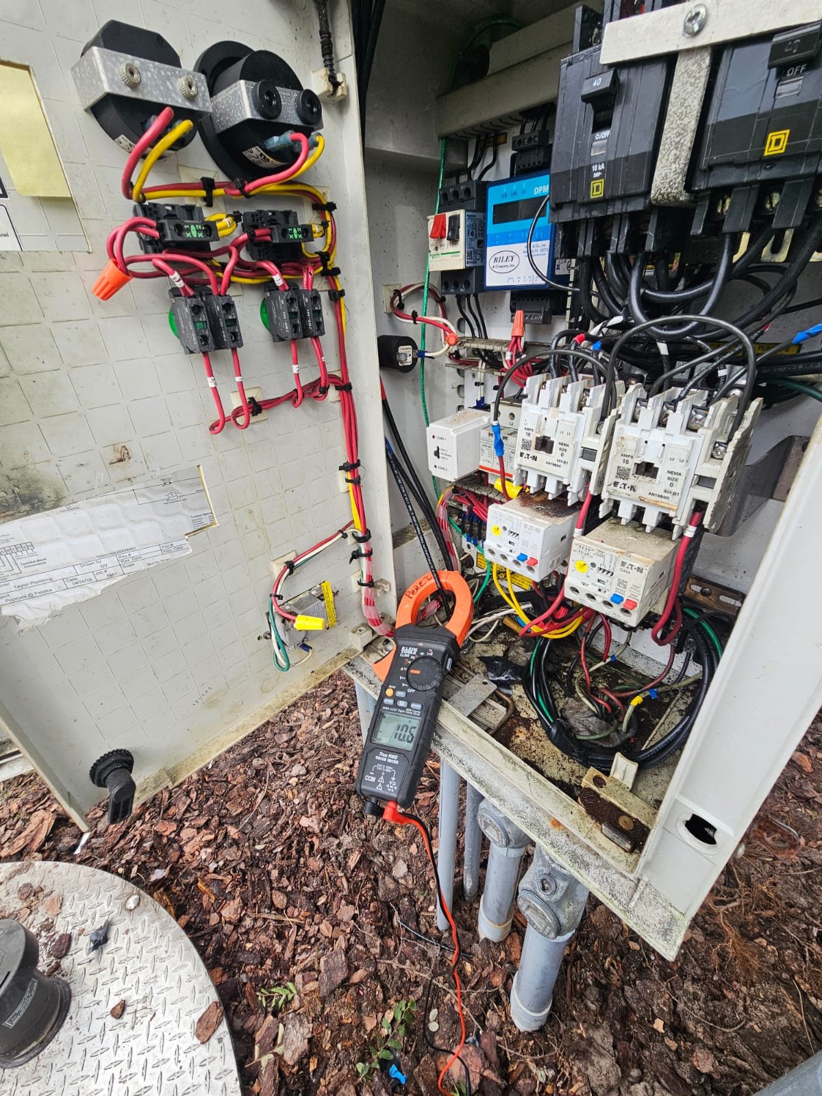 Open electrical panel with various wires, circuit breakers, relays, and a digital multimeter measuring voltage, set outside on ground with gravel surface.