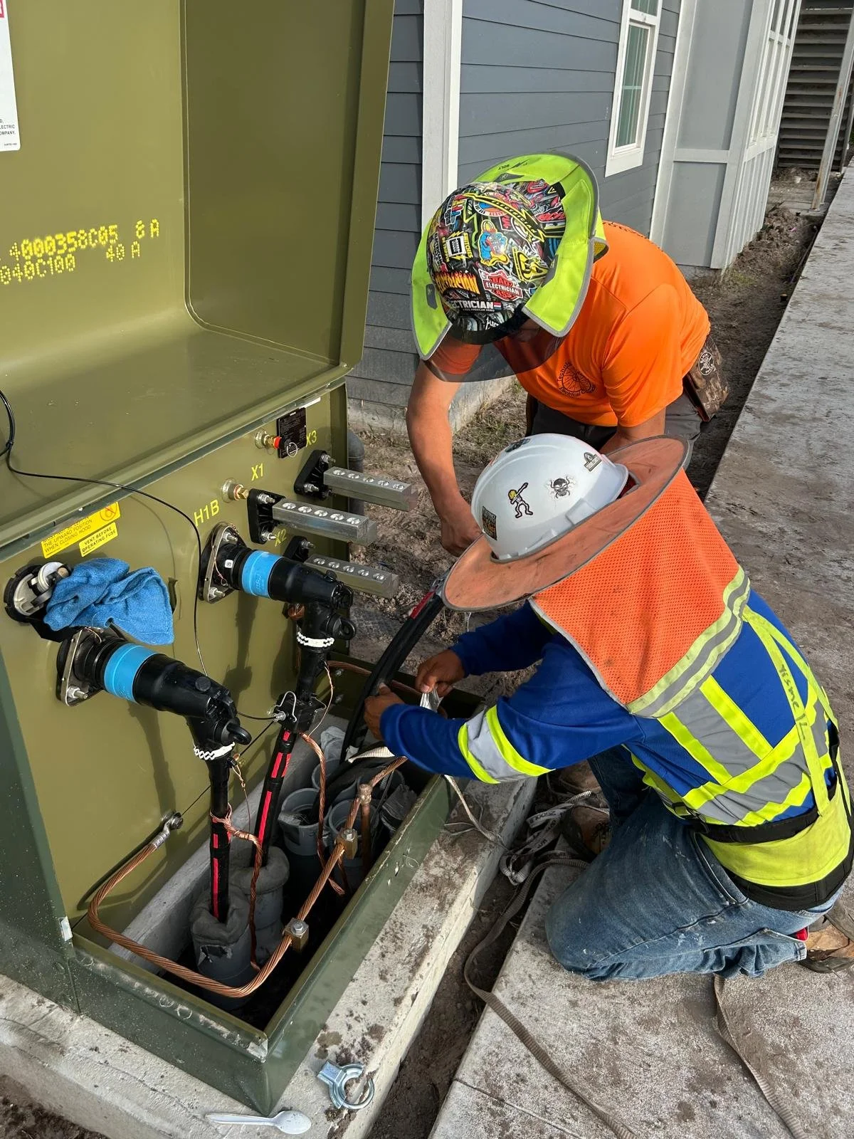 Two construction workers wearing safety helmets and reflective vests working on an electrical or mechanical unit outdoors near a house.