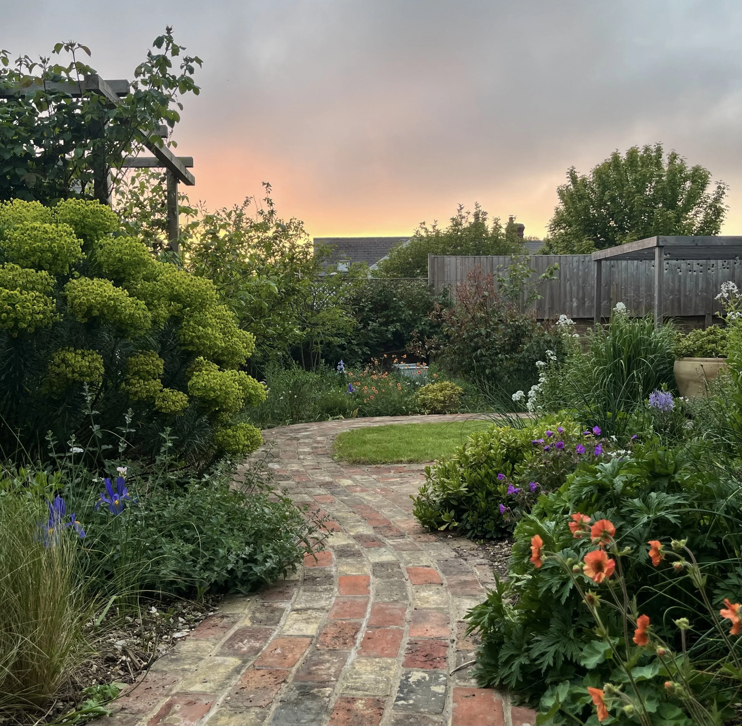 A garden with various green plants and colorful flowers along a brick pathway, with a wooden fence and trees in the background under a cloudy sky during sunset.