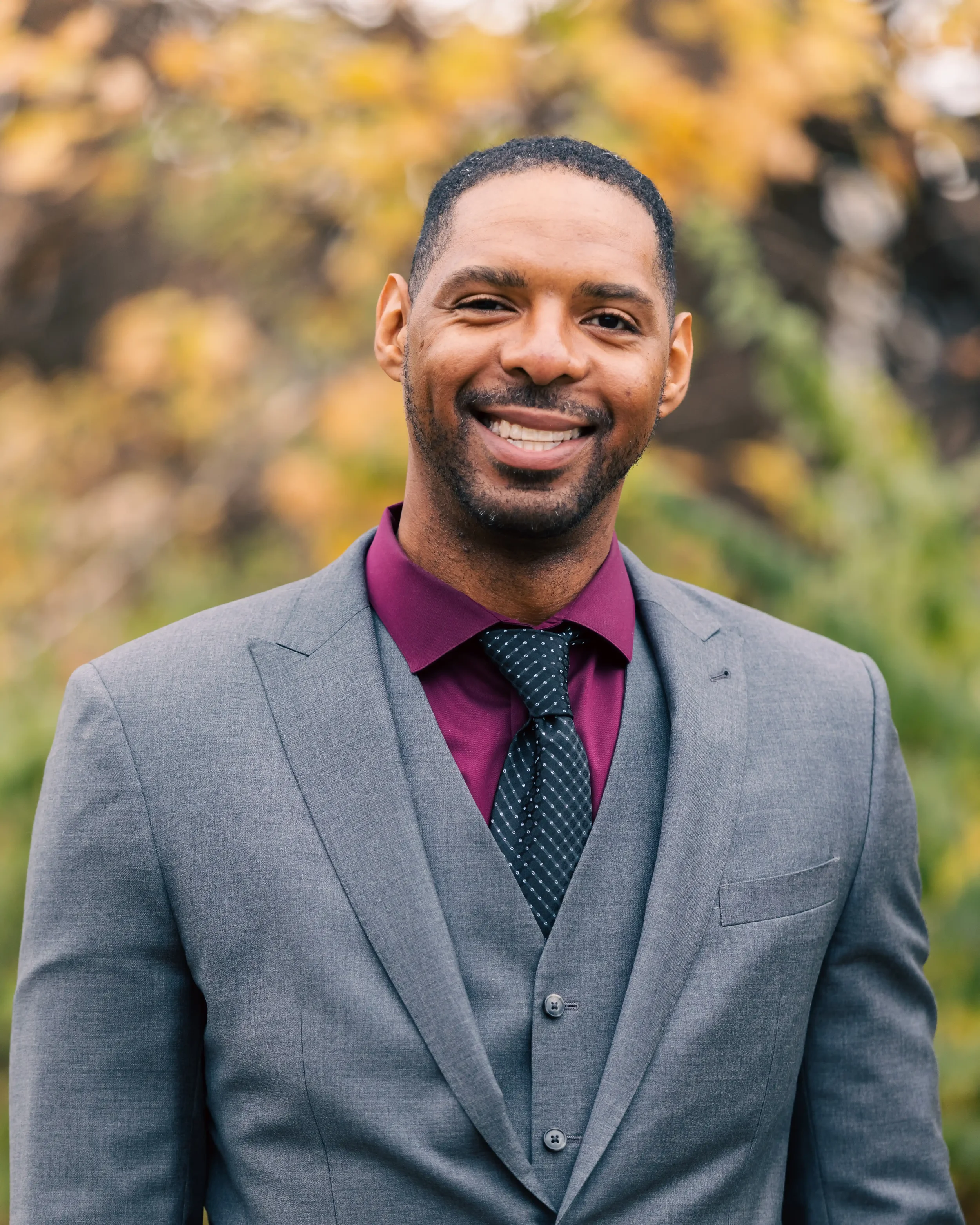 A smiling Zeke in a gray suit and burgundy shirt outdoors with autumn foliage in the background.
