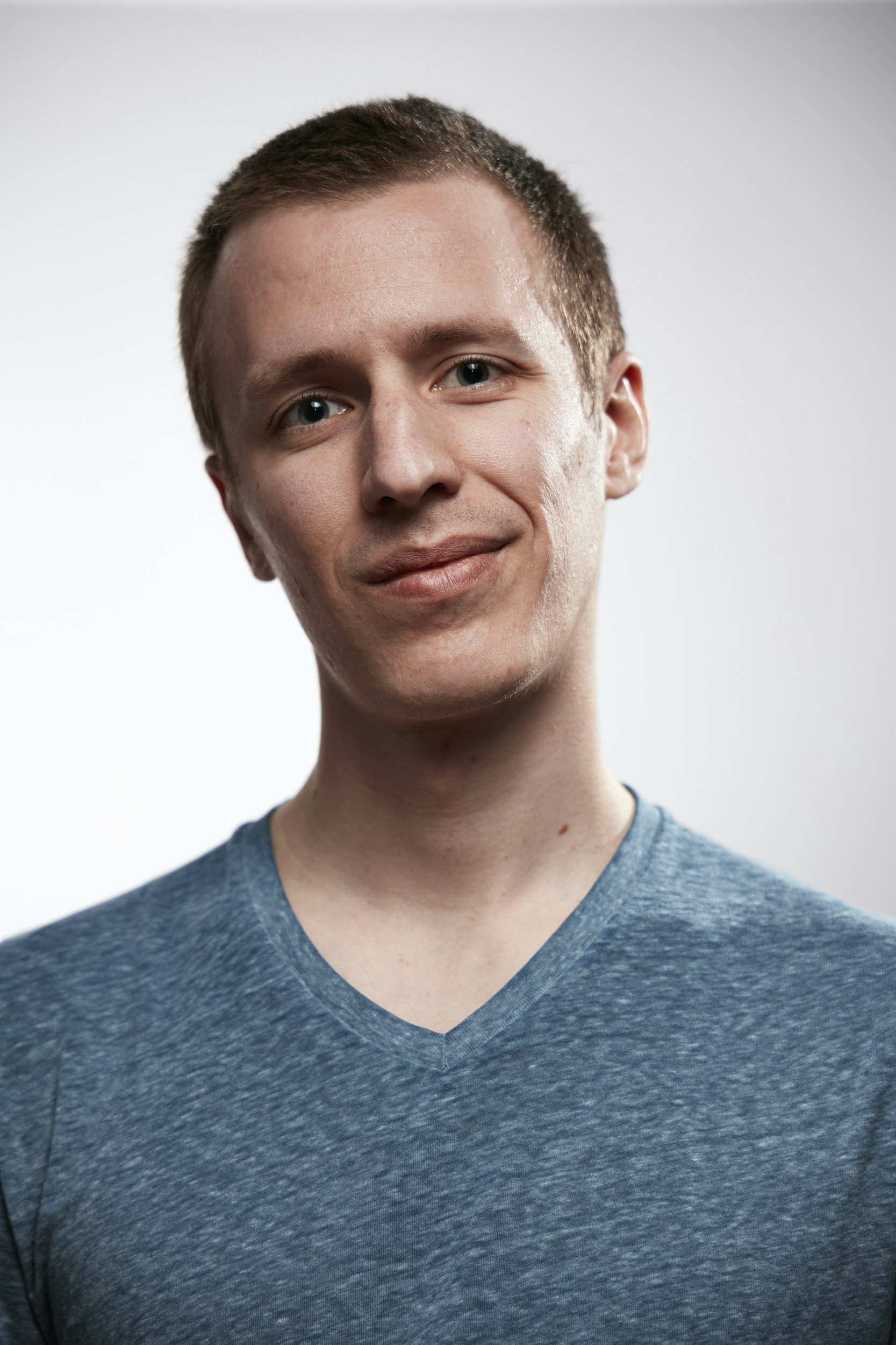 A portrait of a young man with short brown hair and blue eyes, wearing a blue V-neck T-shirt, smiling slightly against a plain white background.