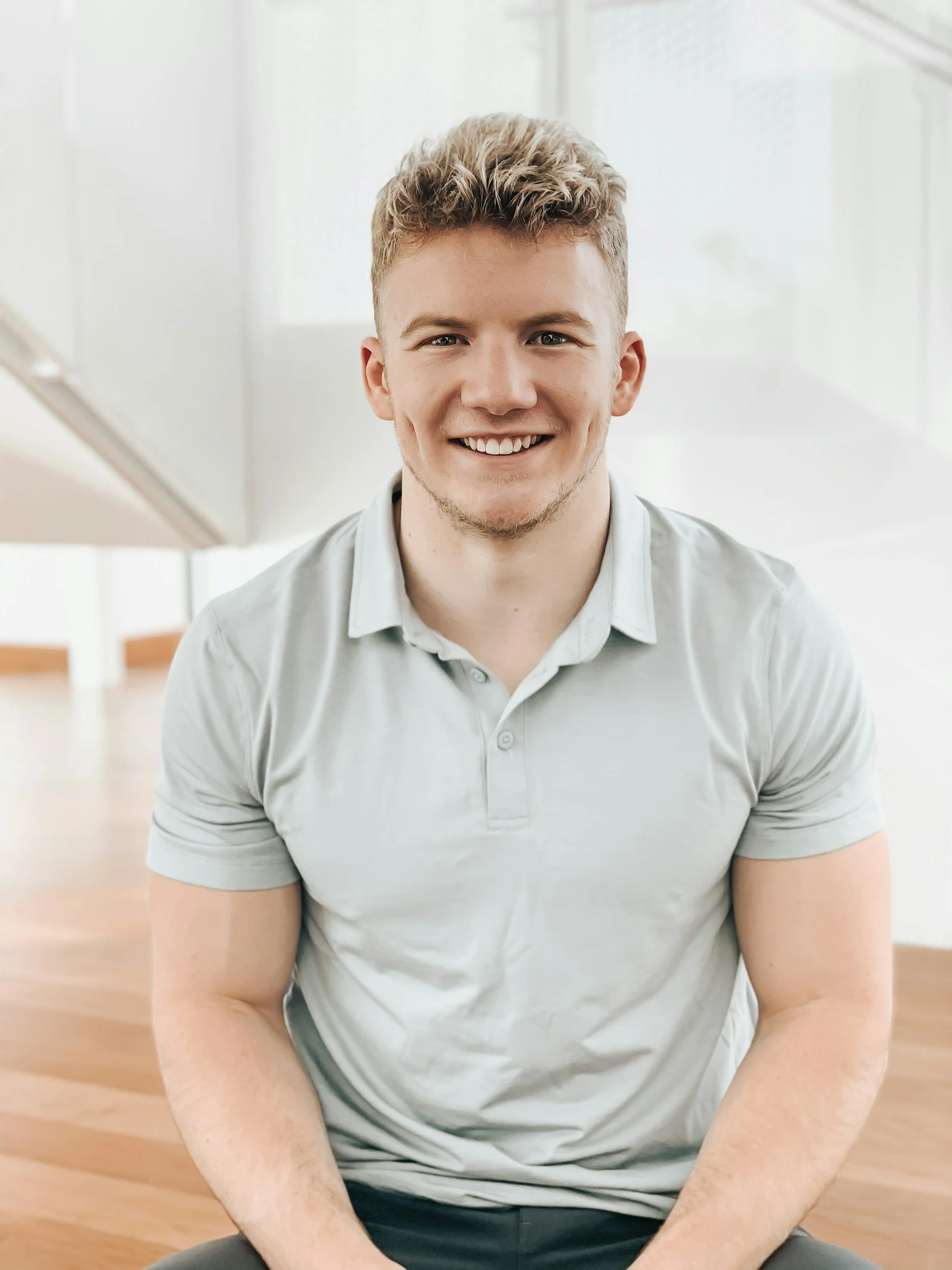 A young man with light brown hair, blue eyes, and a wide smile, sitting indoors on a light wooden floor near a white wall with a window. He wears a light gray polo shirt and appears happy.