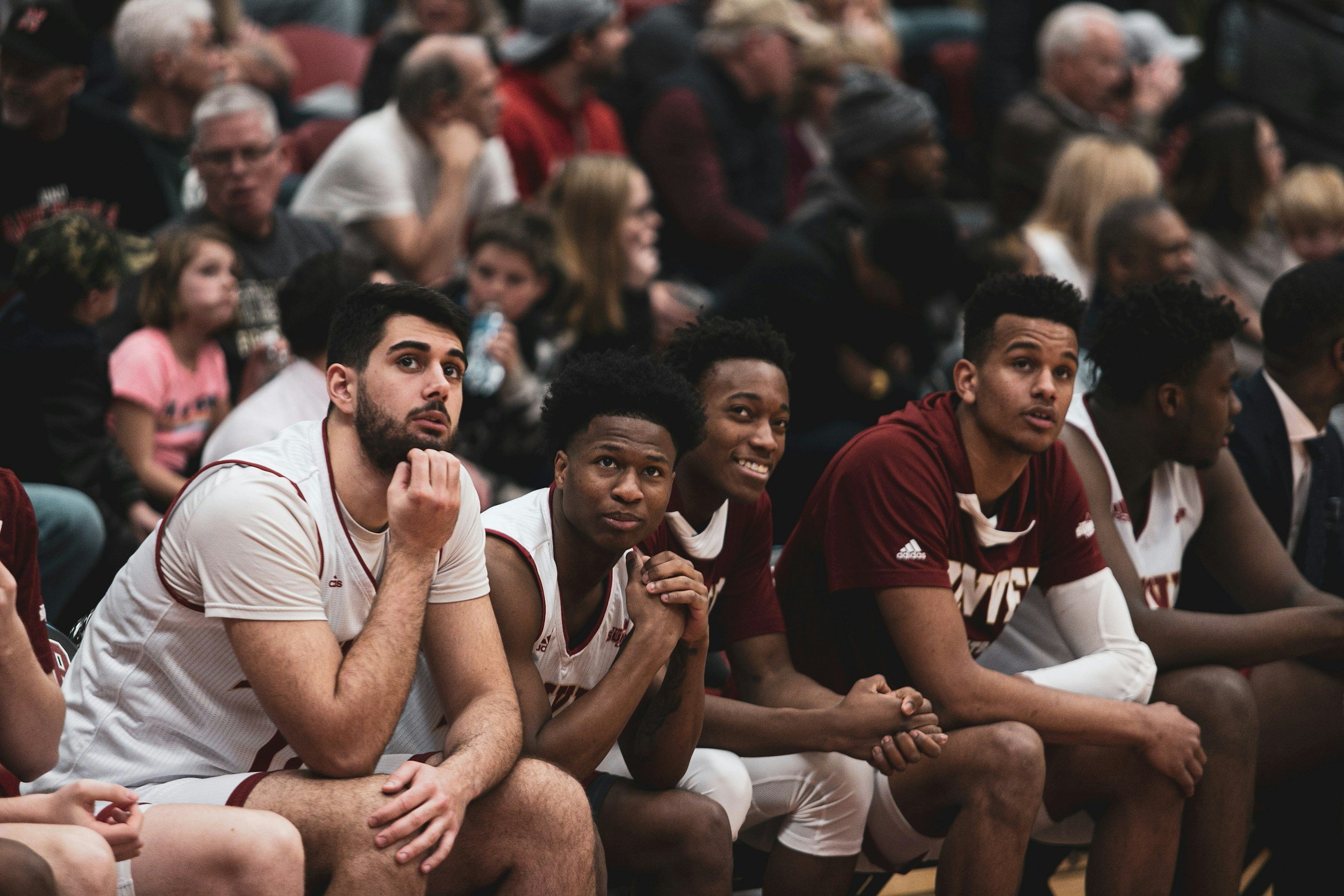 A group of young basketball players sitting courtside, dressed in team uniforms, watching an event with various spectators in the background.