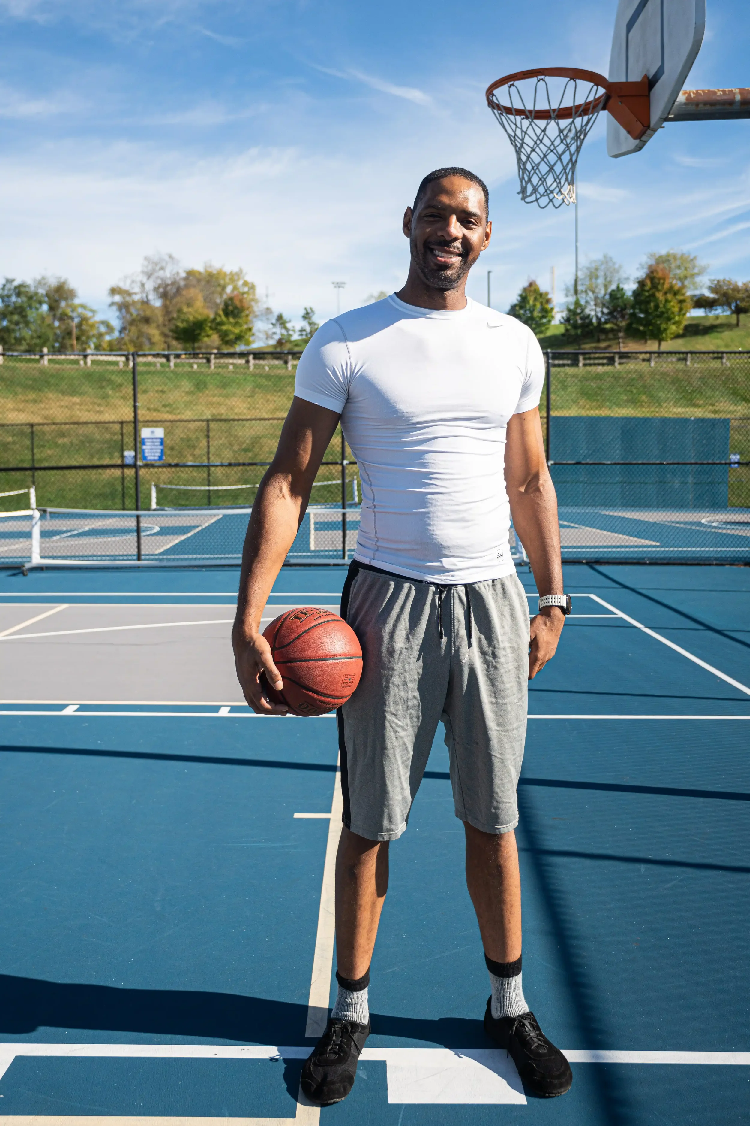 Zeke standing on a basketball court holding a basketball in his right hand. He is smiling, wearing a white athletic shirt, gray shorts, black sneakers, and gray socks. The background shows a blue sky, trees, and a fence surrounding the court.