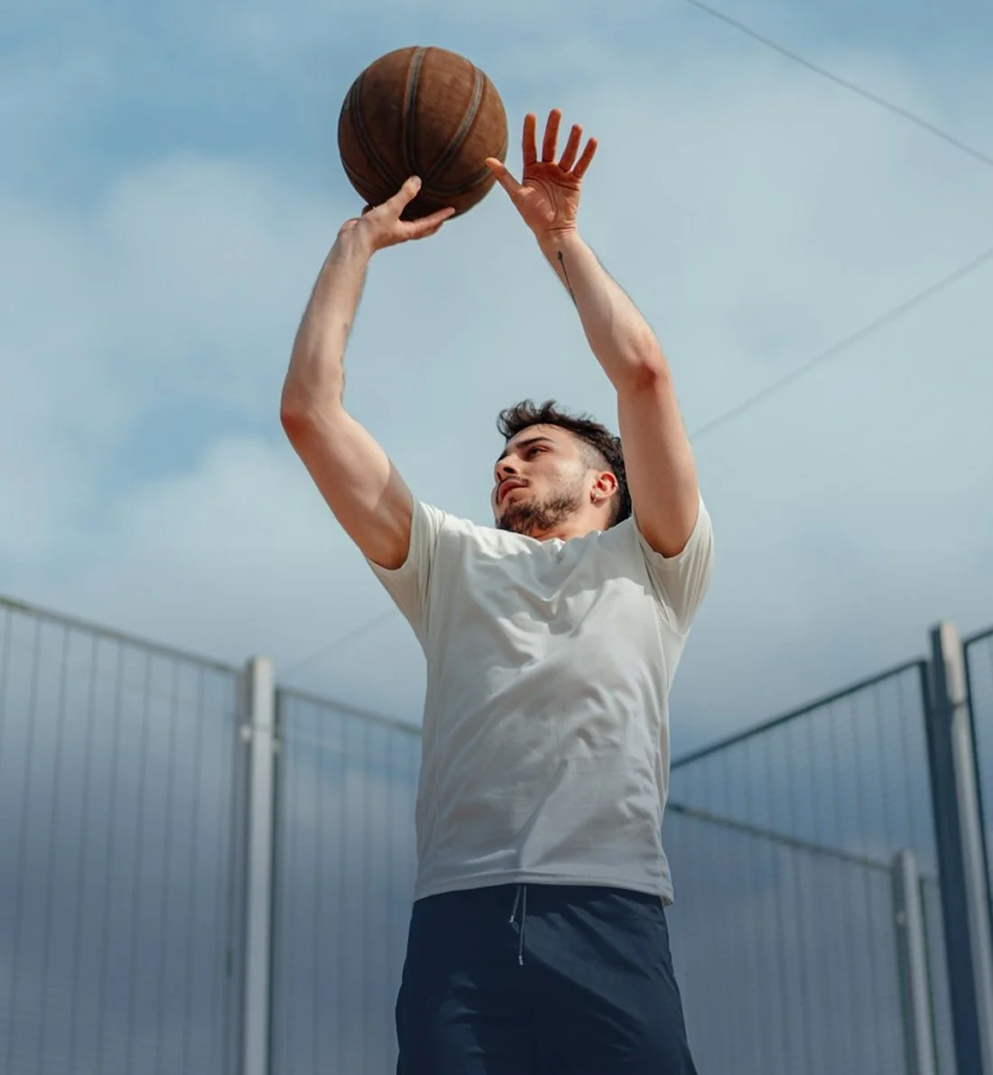 A man in a white shirt and dark shorts shooting a basketball on an outdoor court.