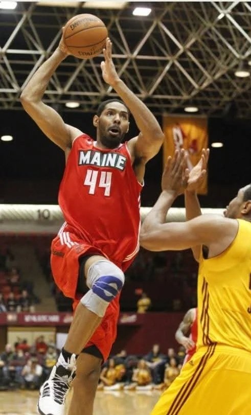 A basketball player from Maine, wearing jersey number 44 in red, is jumping with a basketball in his hands, attempting a shot, while a defender in yellow tries to block him in an indoor basketball court.