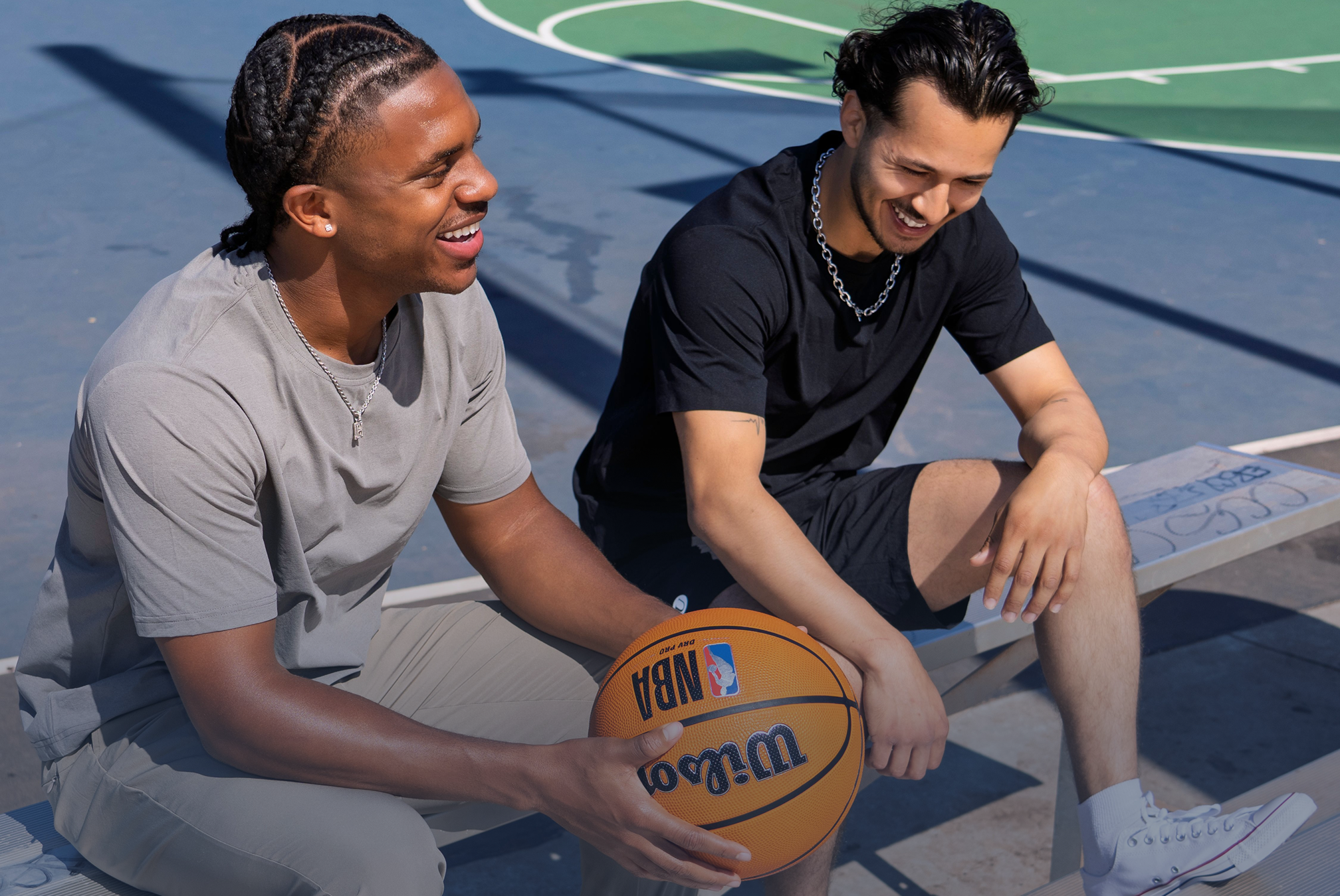Two young men sitting on a bench at an outdoor basketball court, smiling and engaging in conversation. One is holding a Wilson NBA basketball, and the court has a blue and green surface with white lines.