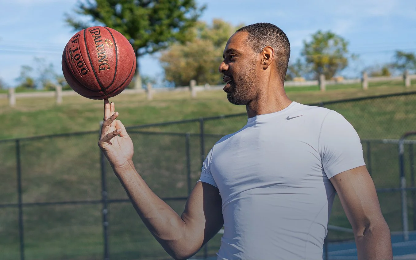 Zeke in a white athletic shirt spinning a basketball on his finger on an outdoor basketball court.