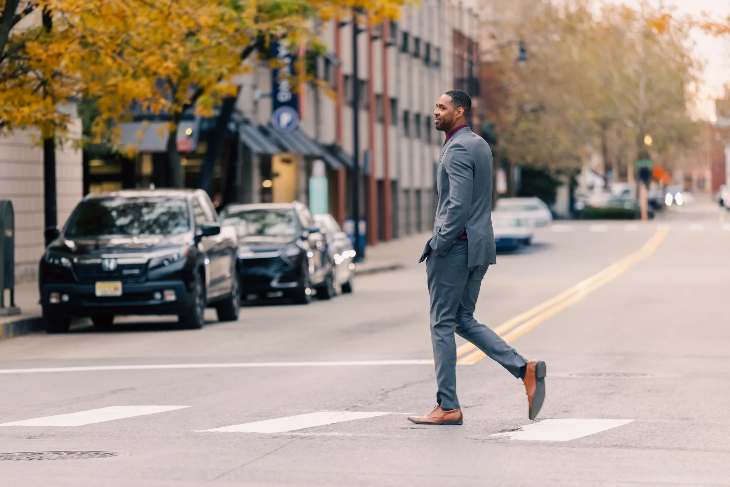 Zeke in a gray suit and brown shoes crossing a street during daytime with trees showcasing fall foliage in the background.