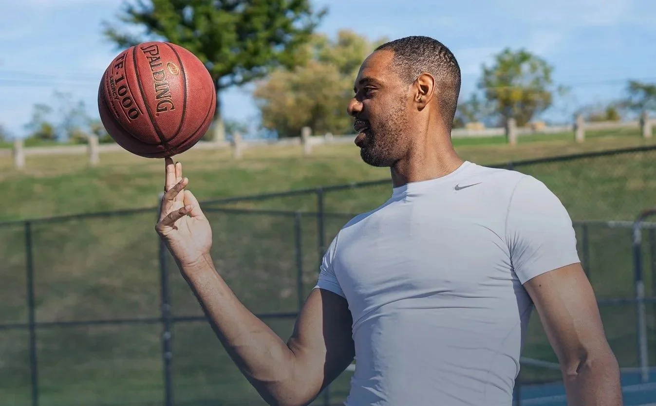 Zeke in a white athletic shirt spins a basketball on his finger outdoors on a sunny day, with a grassy field and trees in the background.