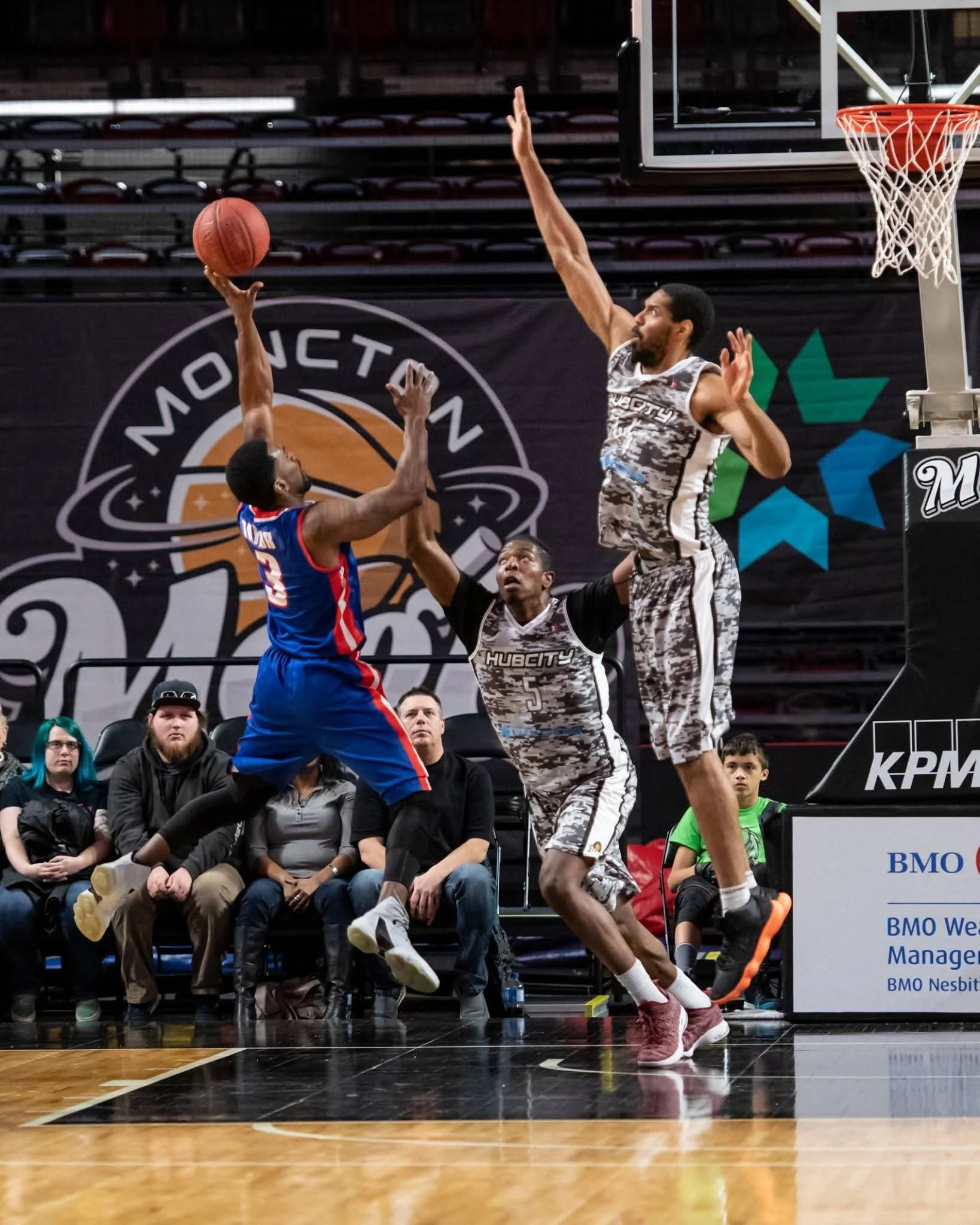 Two basketball players leaping for a rebound during a game. The player in the blue and red uniform is reaching for the ball, while the player in the camouflage-patterned uniform is trying to block him. Spectators watch from the stands in the background.