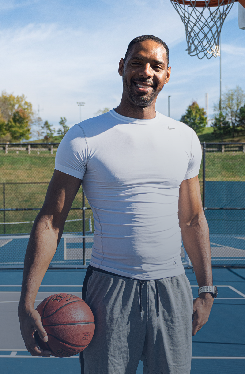 Zeke in a white athletic shirt and gray shorts holding a basketball on an outdoor basketball court.