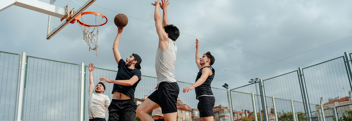 Four young men playing basketball outdoors on a fenced court during daytime, with one attempting a shot near the hoop and the others watching or defending.