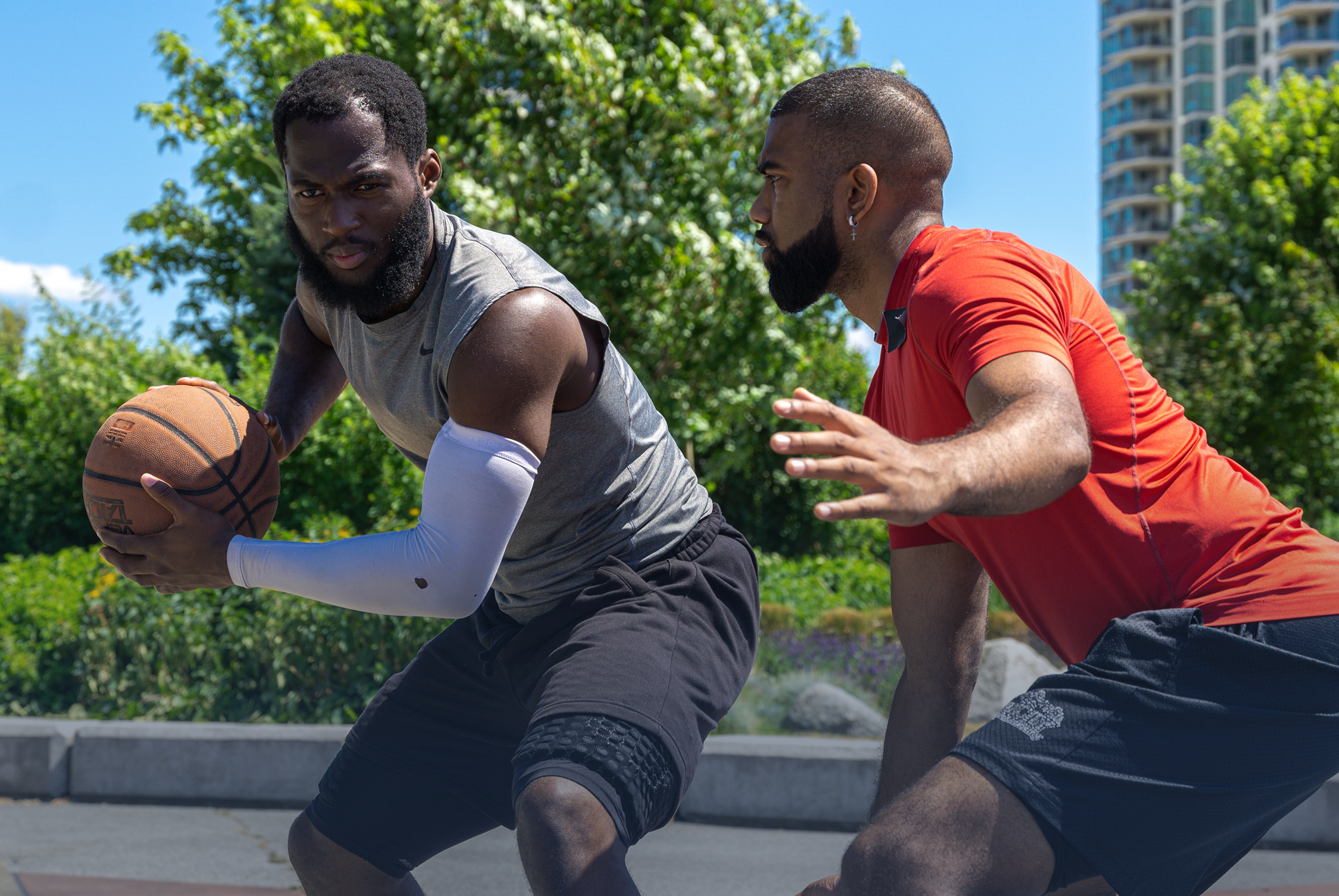 Two men playing basketball outdoors on a sunny day, with trees and a high-rise building in the background. One man in a gray sleeveless shirt is holding a basketball, while the other man in a red shirt is defending.