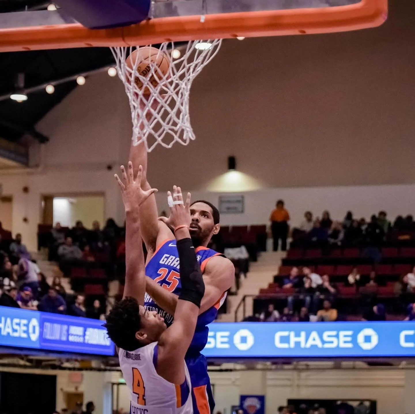 A basketball player in a blue jersey, number 25, attempting a shot at the basket while an opponent in a white jersey, number 4, reaches up defensively in a basketball game.