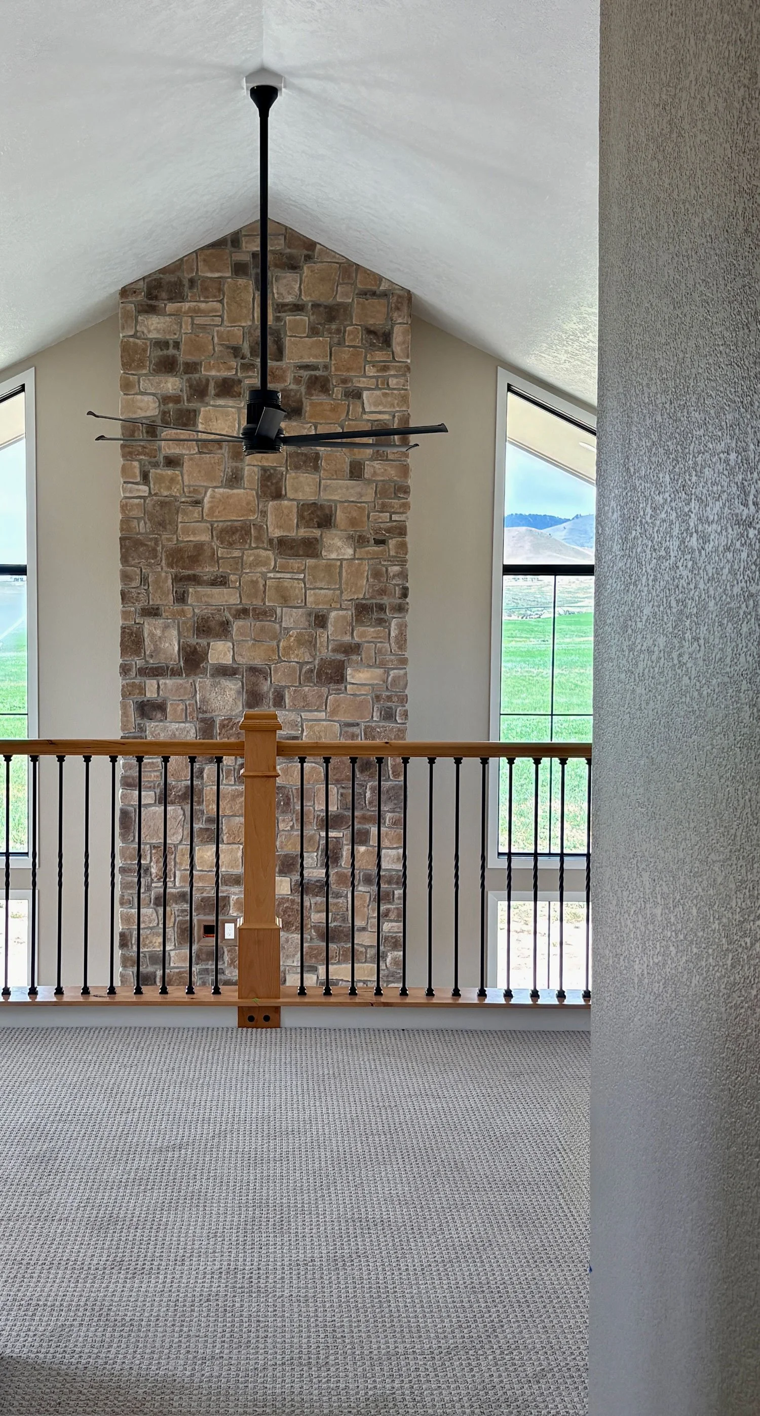 Interior view of a home with a high vaulted ceiling, a stone chimney, large windows showing a scenic landscape, and a modern ceiling fan.
