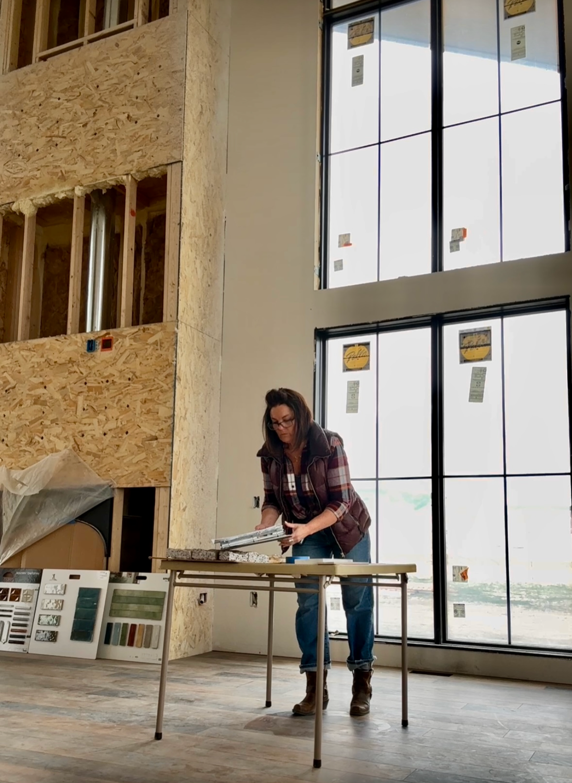 A woman in construction gear inspecting paint samples at a building site with large windows and unfinished walls.