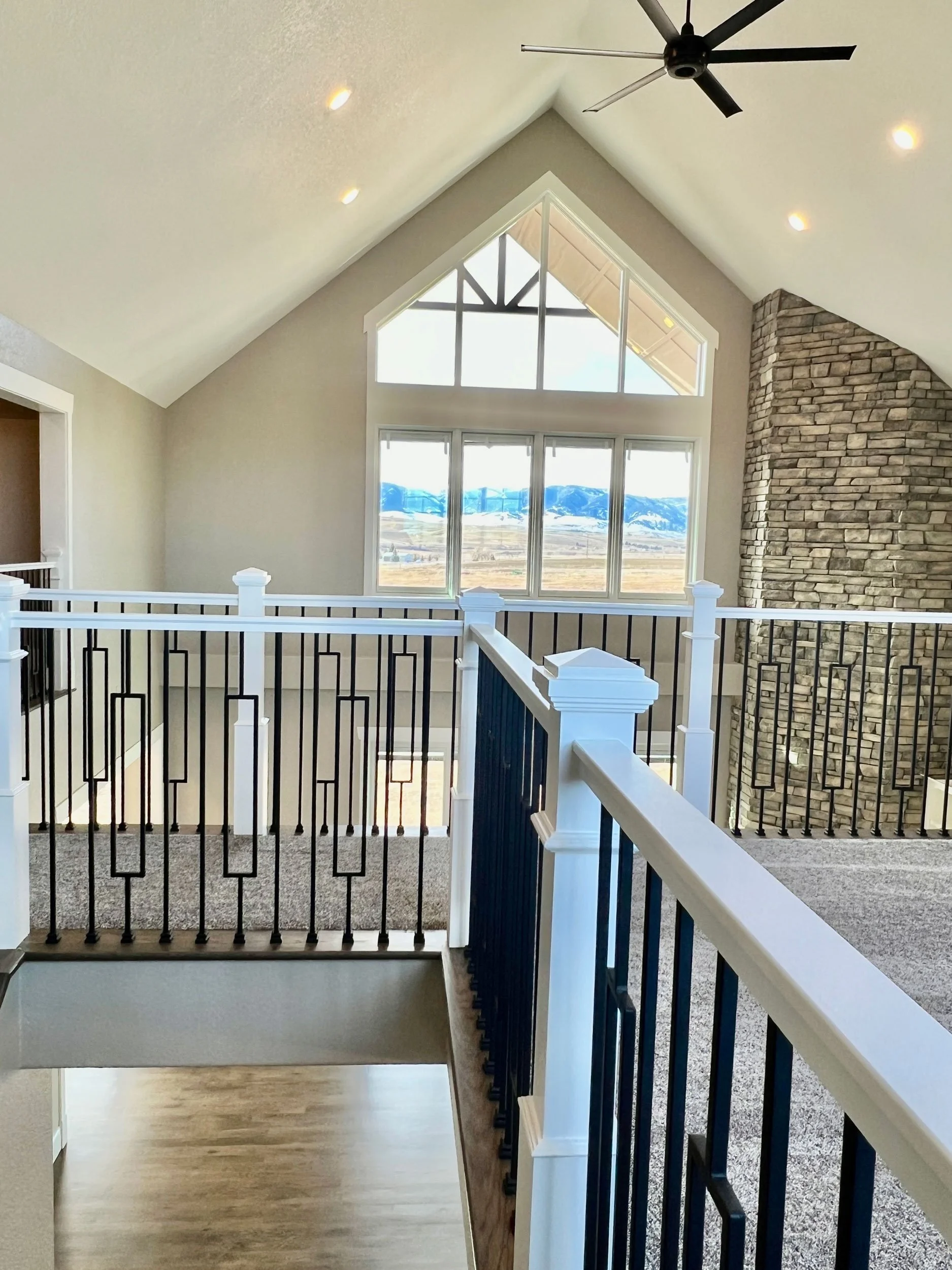 Interior view of a modern home with large windows showing mountains in the background, featuring a black ceiling fan, white walls, hardwood and carpeted floors, a stone chimney, and a decorative black and white railing.