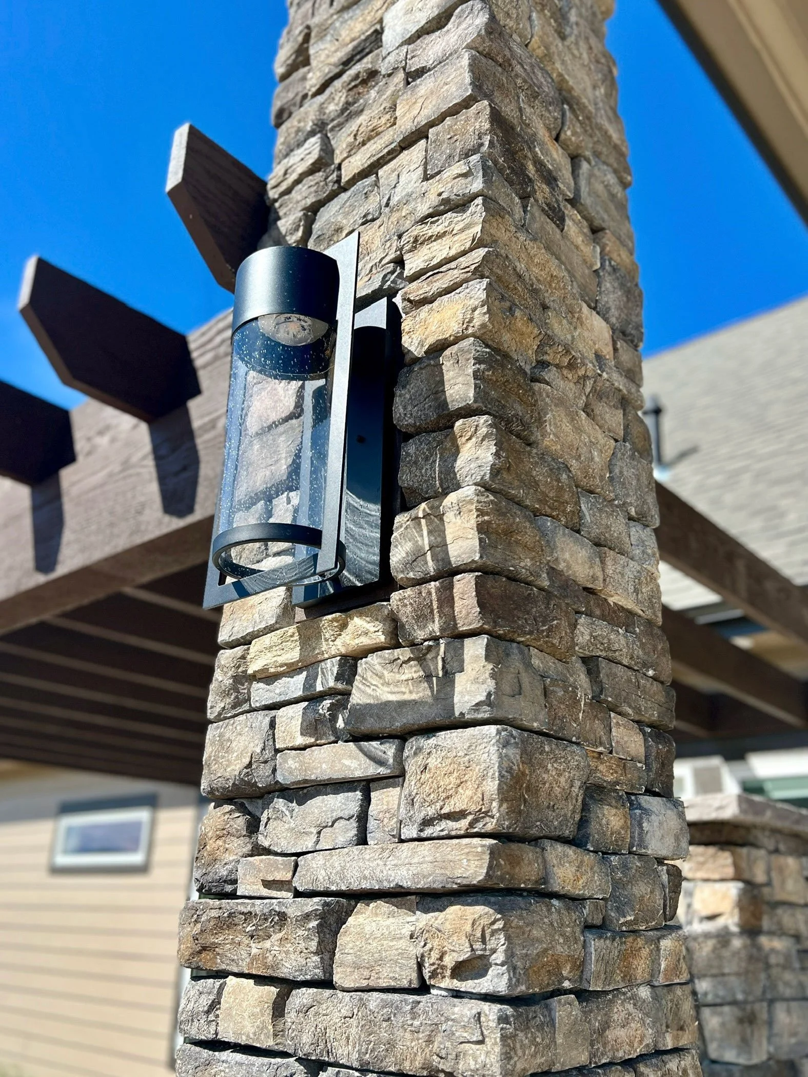 Close-up of a stone pillar with a modern wall-mounted outdoor light fixture against a clear blue sky and part of a residential building in the background.