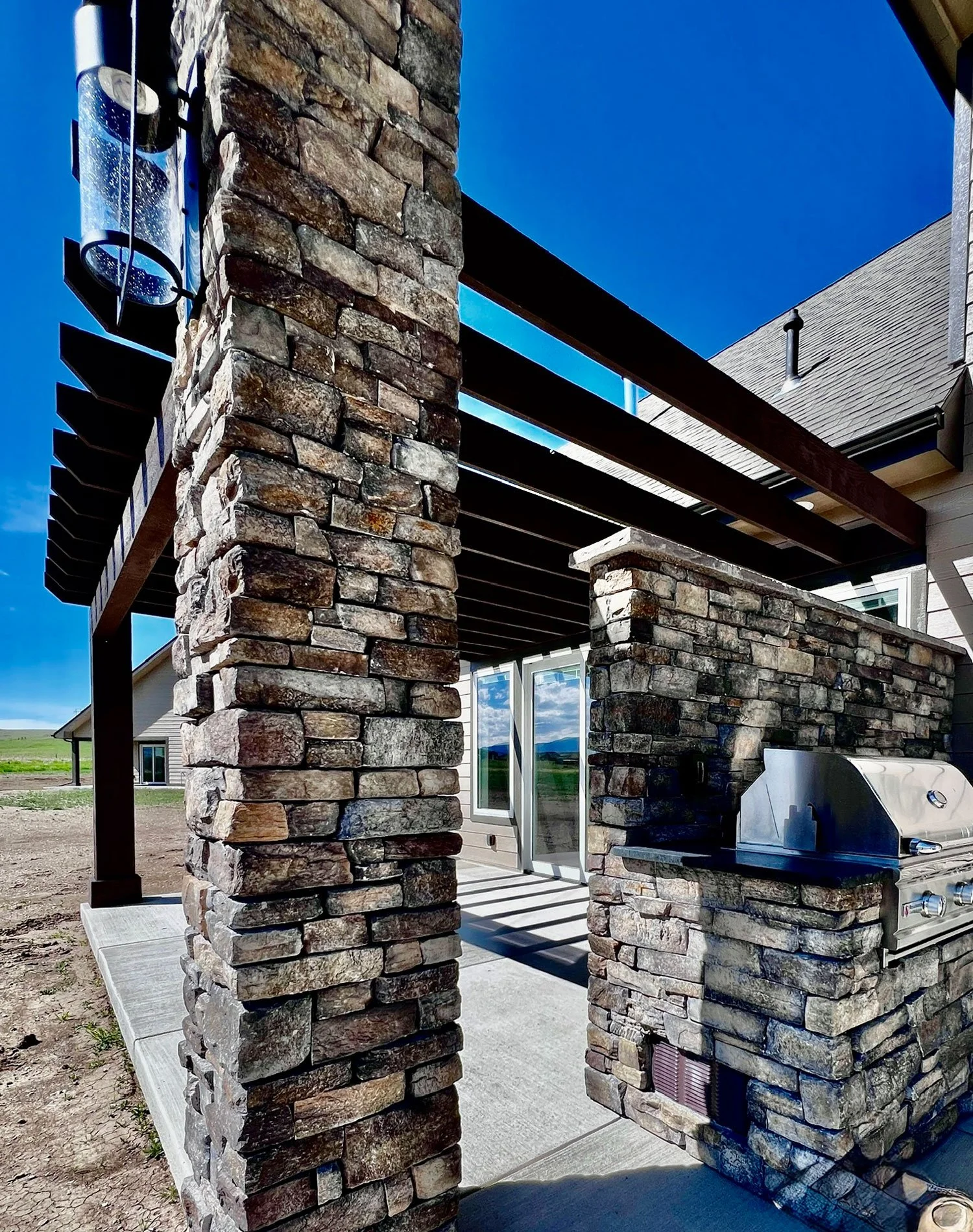A close-up of a stone pillar with a built-in outdoor grill, part of a patio area under a wooden pergola, with another building and a grassy field in the background.