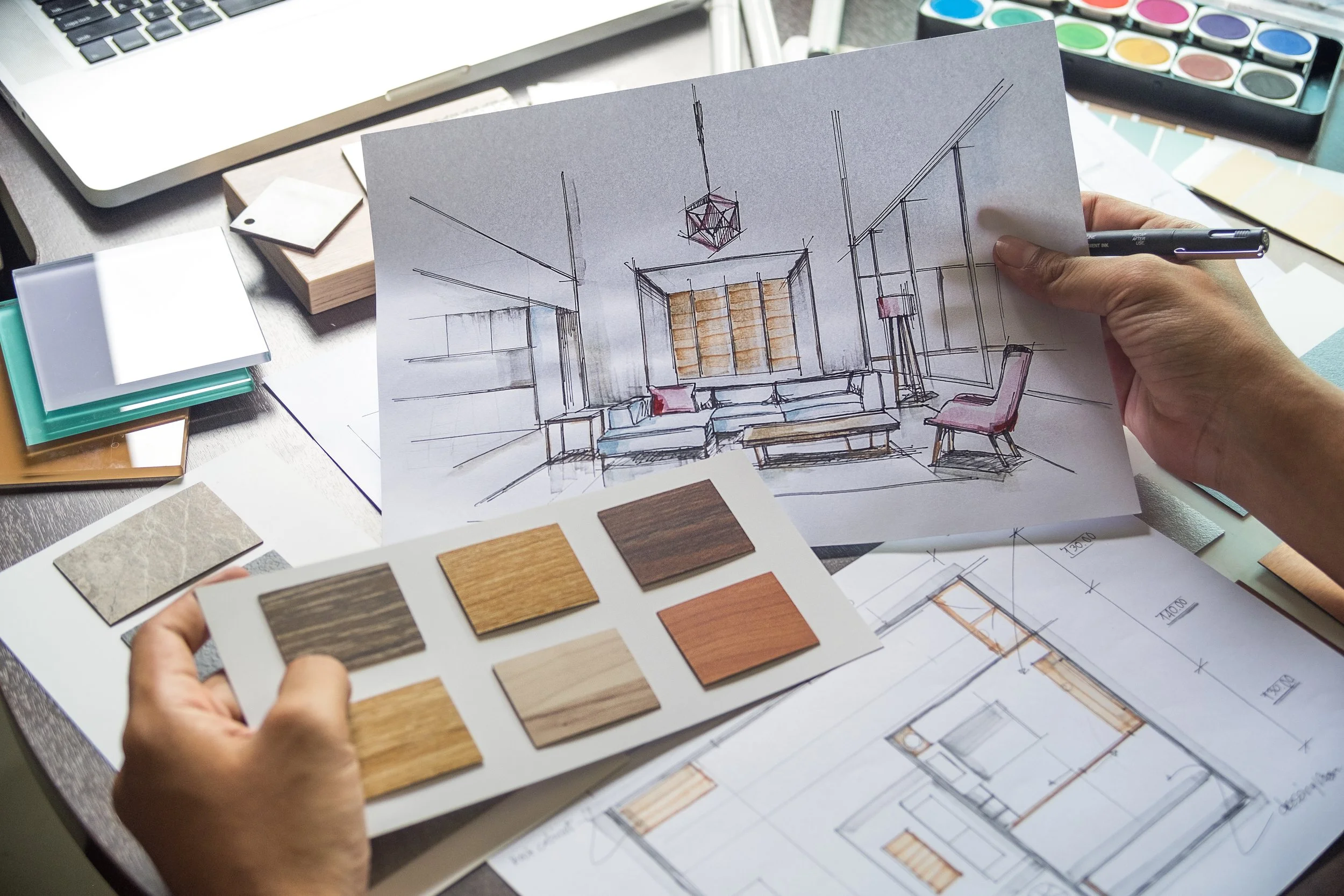 A person holds a sketch of a modern living room interior design with wooden furniture and a geometric ceiling light fixture. The workspace includes color sample swatches, architectural plans, a laptop, and watercolor paints.