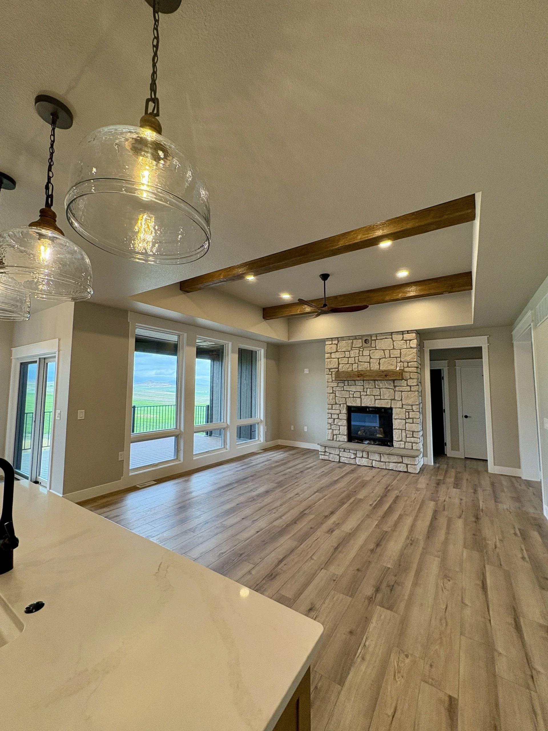 A living room with large windows, a stone fireplace, wooden beams on the ceiling, hardwood flooring, and a view of green fields outside.