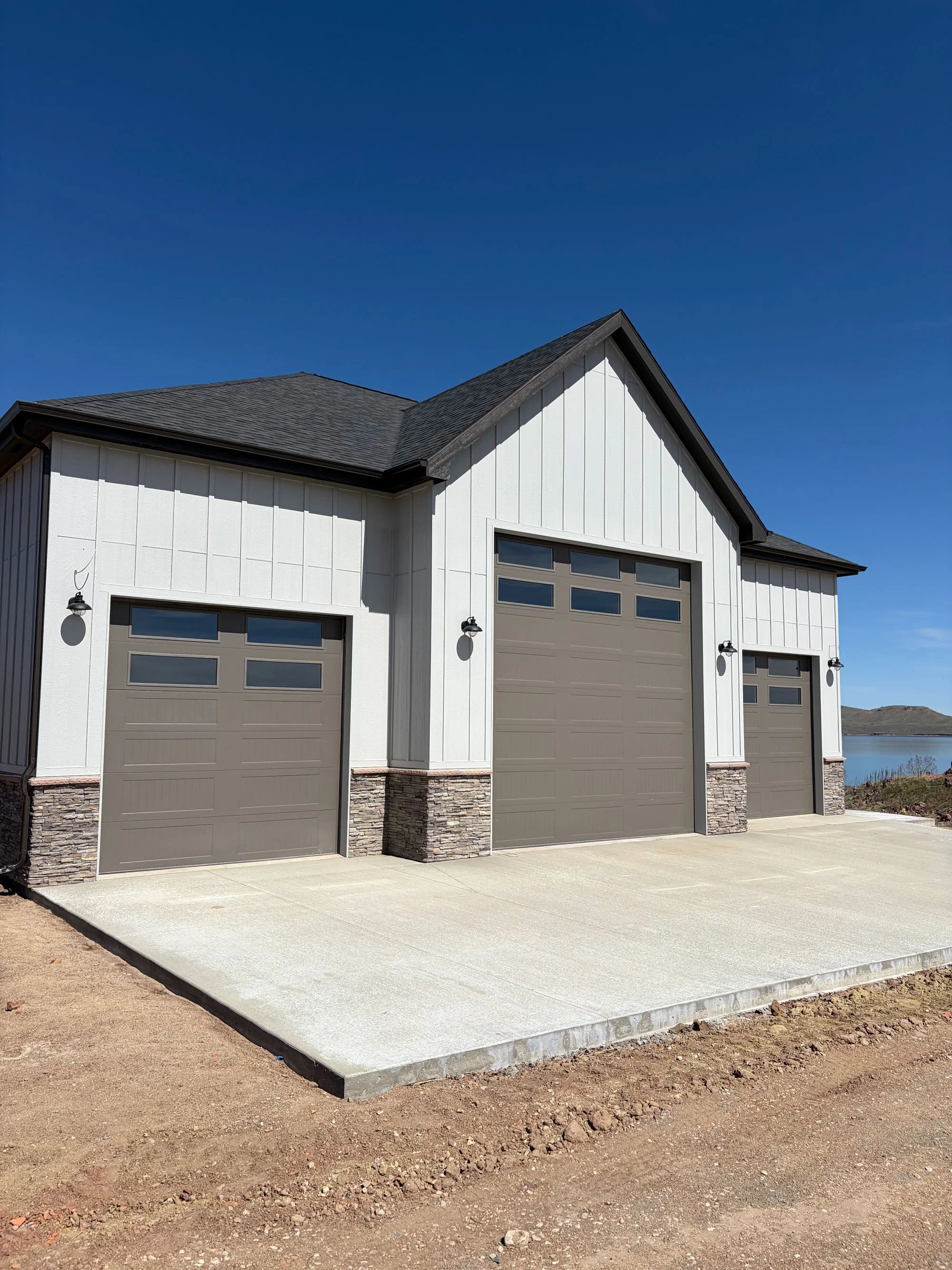 Newly constructed garage with a concrete driveway, white siding, gray doors, stone accents at the base, and black outdoor lights, set against a mountain and water background under a clear blue sky.