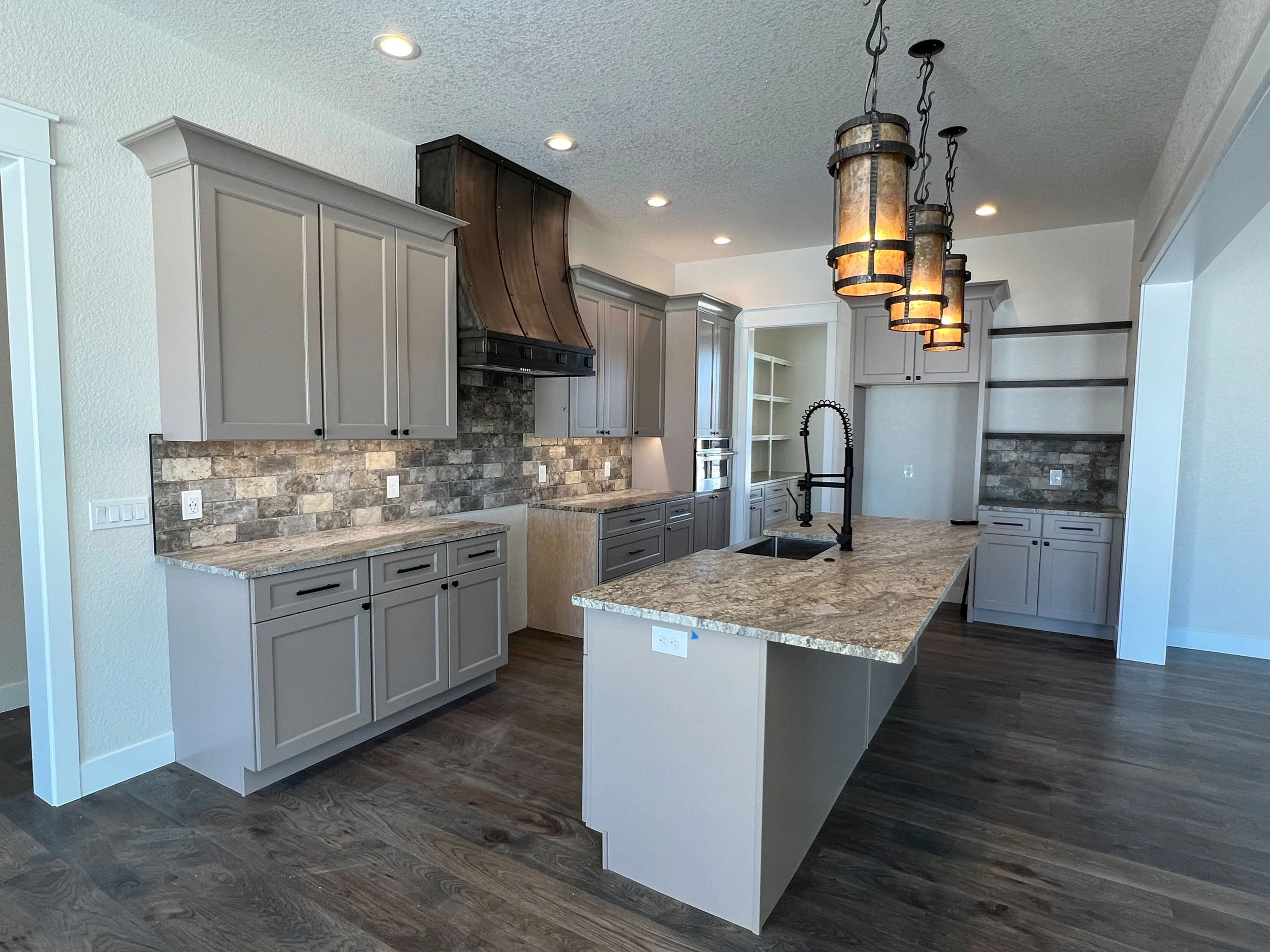 Modern kitchen with beige cabinets, brick backsplash, kitchen island with granite top, pendant lights, stainless steel oven, black sink and faucet, and dark wood flooring.