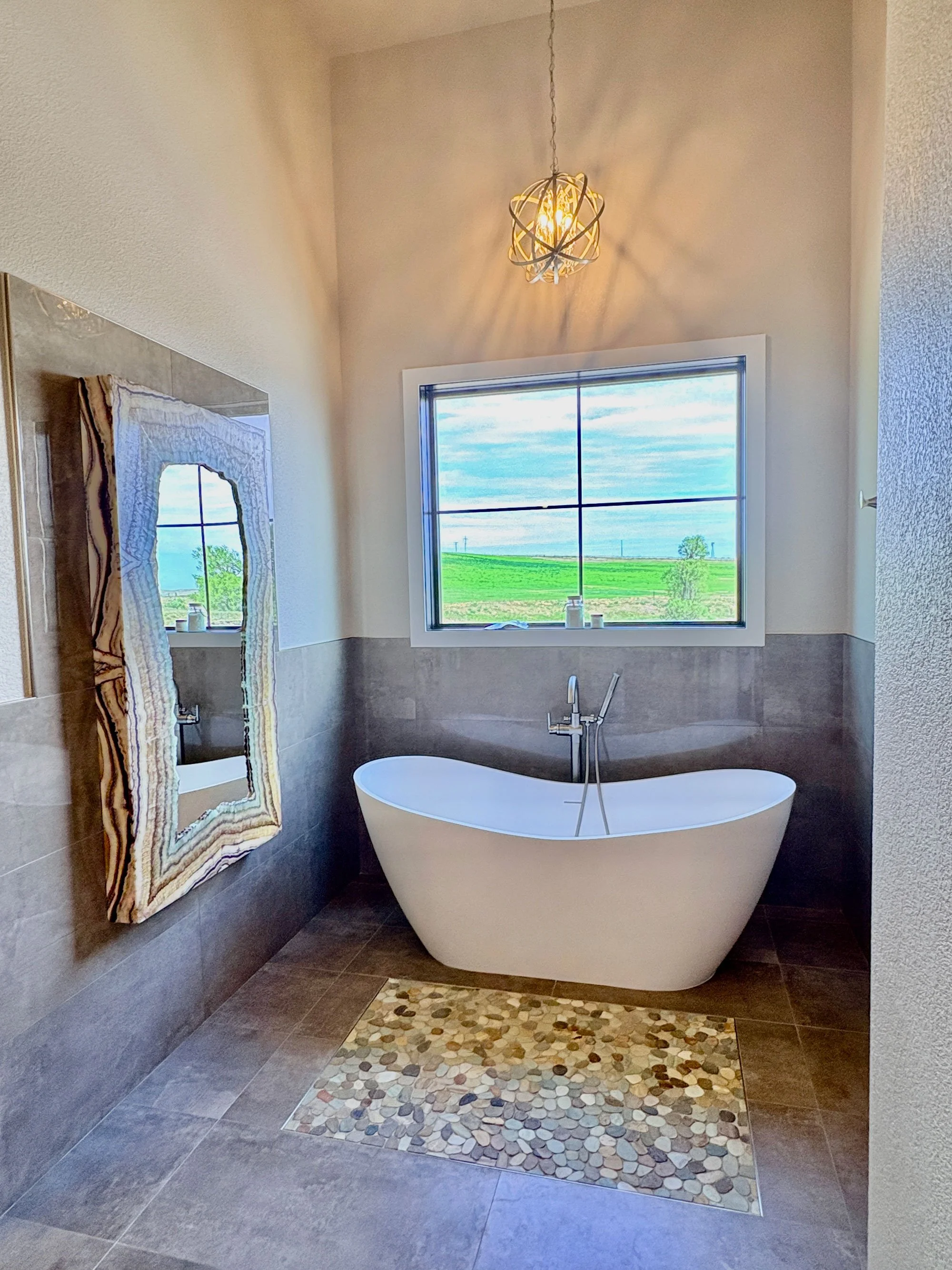 A bathroom with a freestanding bathtub under a window with a view of green fields and trees outside. There is a pebble stone rug in front of the tub, an ornate mirror on the wall, and a decorative light fixture hanging from the ceiling.