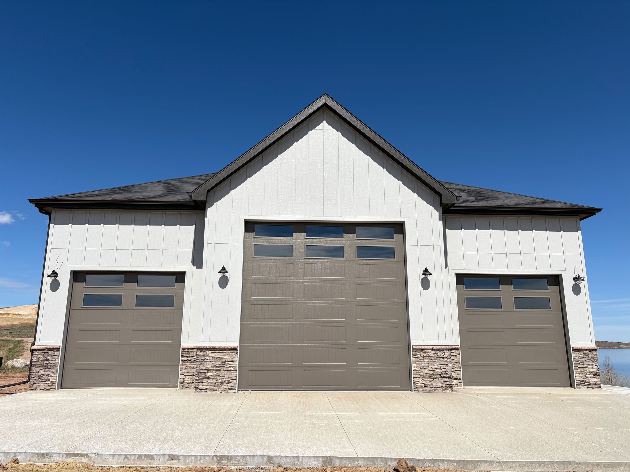Front view of a modern house with two garage doors, white siding, stone accents, and a peaked roof, under a clear blue sky.