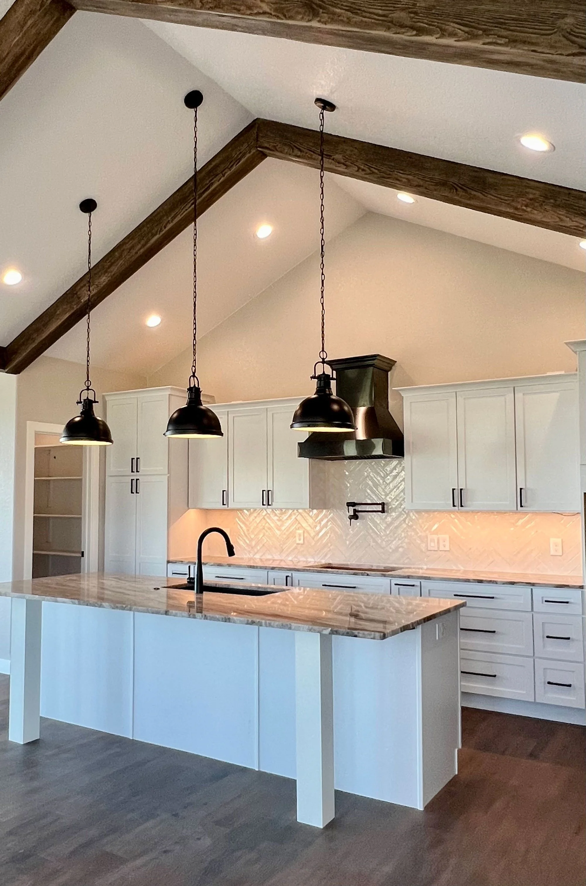 Modern kitchen with white cabinets, a marble island, a black sink faucet, a stainless steel range hood, and black pendant lights hanging from a ceiling with exposed wooden beams.