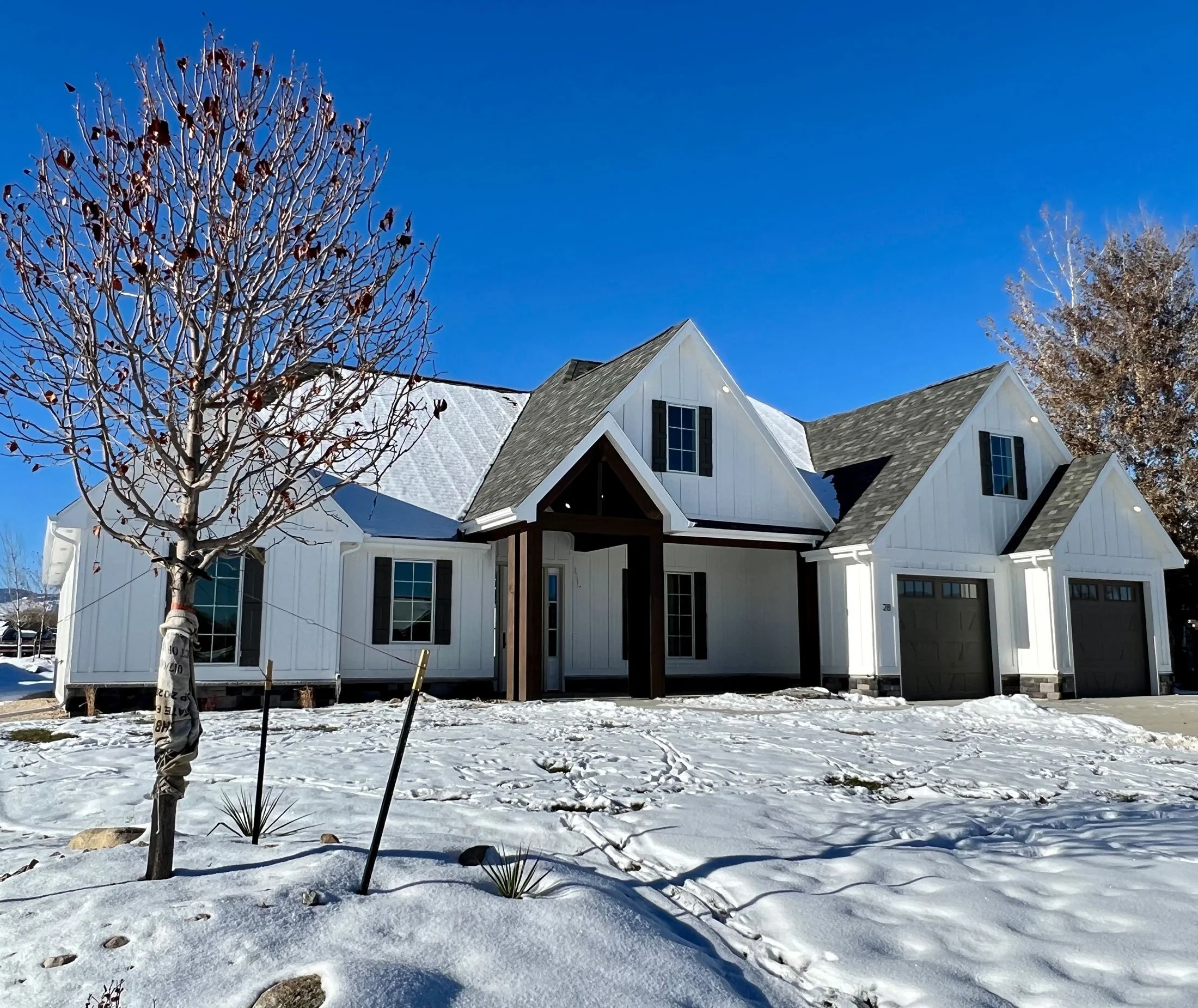 A white house with multiple peaked roofs, black shutters, and several windows, set against a clear blue sky. There are leafless trees and snow on the ground in the foreground.