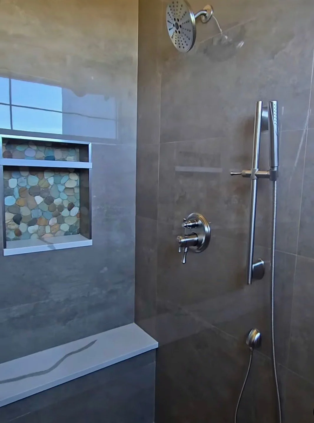 Shower with a rain shower head, a handheld shower Wand, a chrome faucet, a built-in wall niche with multi-colored stones, and neutral-colored tiled walls.