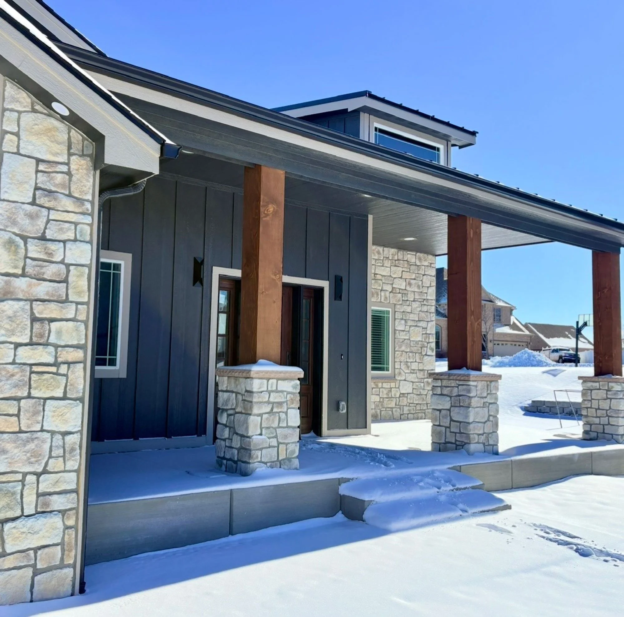 A modern house exterior with stone and dark siding, wooden beams, and a sloped metal roof, surrounded by snow on the ground under a clear blue sky.