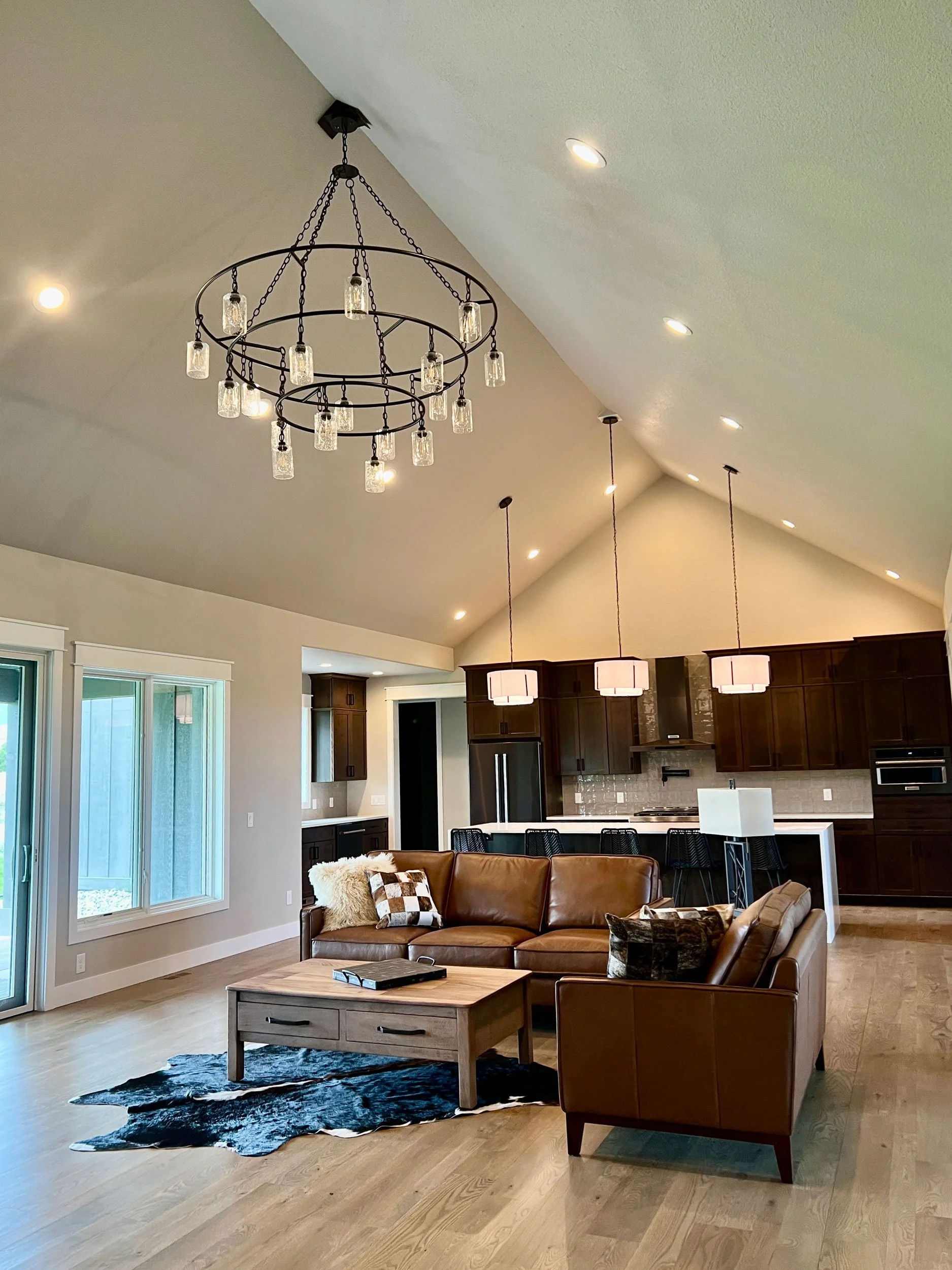 Open-concept living room and kitchen with brown leather sofa, wooden coffee table, cowhide rug, light-colored hardwood floors, and modern lighting fixtures including a chandelier and pendant lights. Large windows provide natural light.