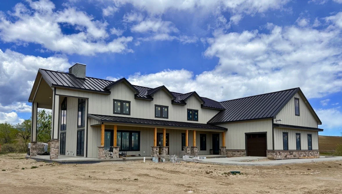 Newly constructed two-story house with a modern design, metal roof, and large windows, situated on a bare plot of land under a partly cloudy sky.